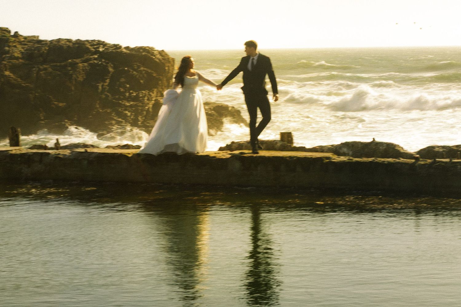 Bride and groom holding hands on rocky coastal cliffs at golden sunset with ocean waves crashing behind them.