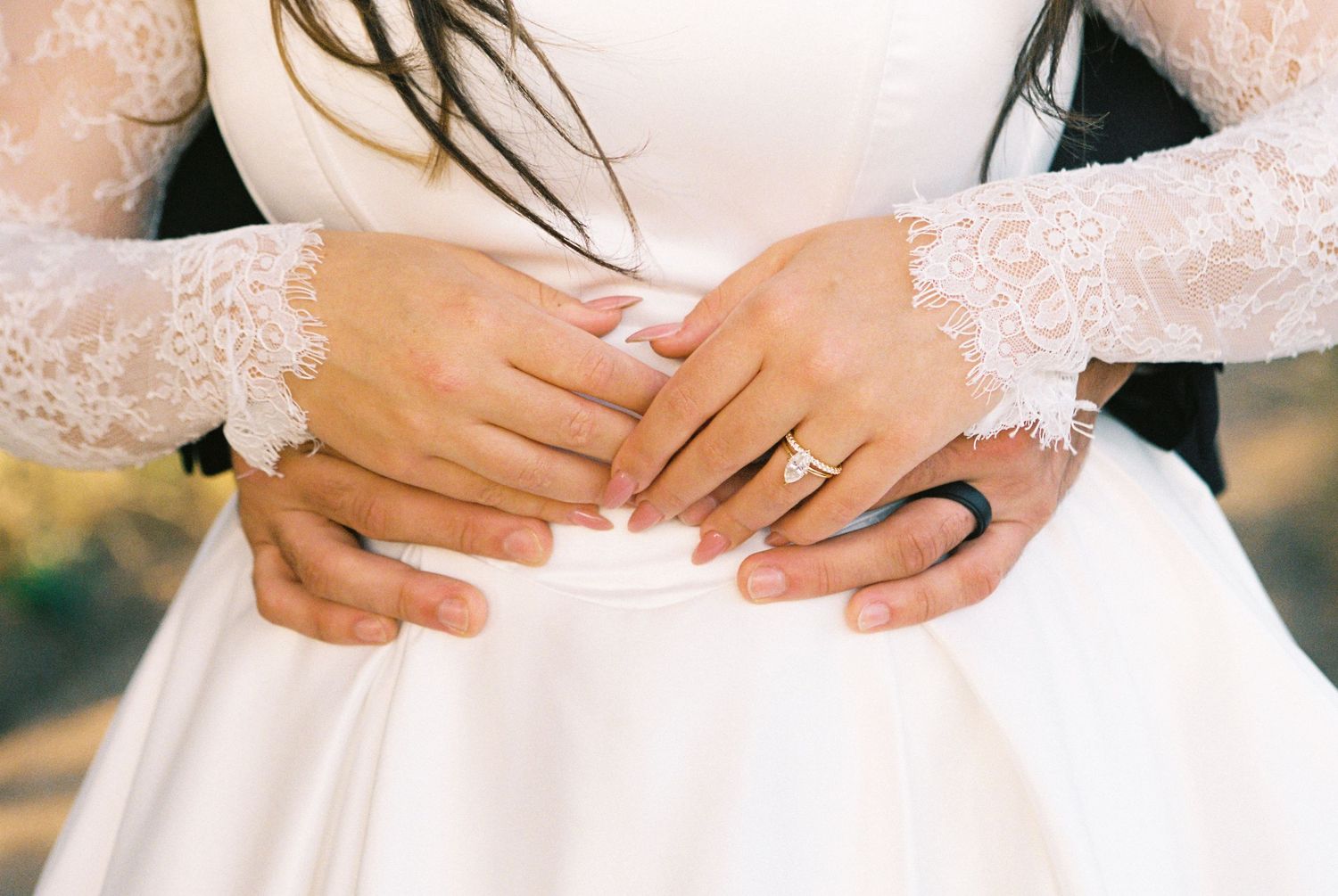 Bride and groom holding hands showing wedding ring on white lace bridal gown at outdoor wedding ceremony.