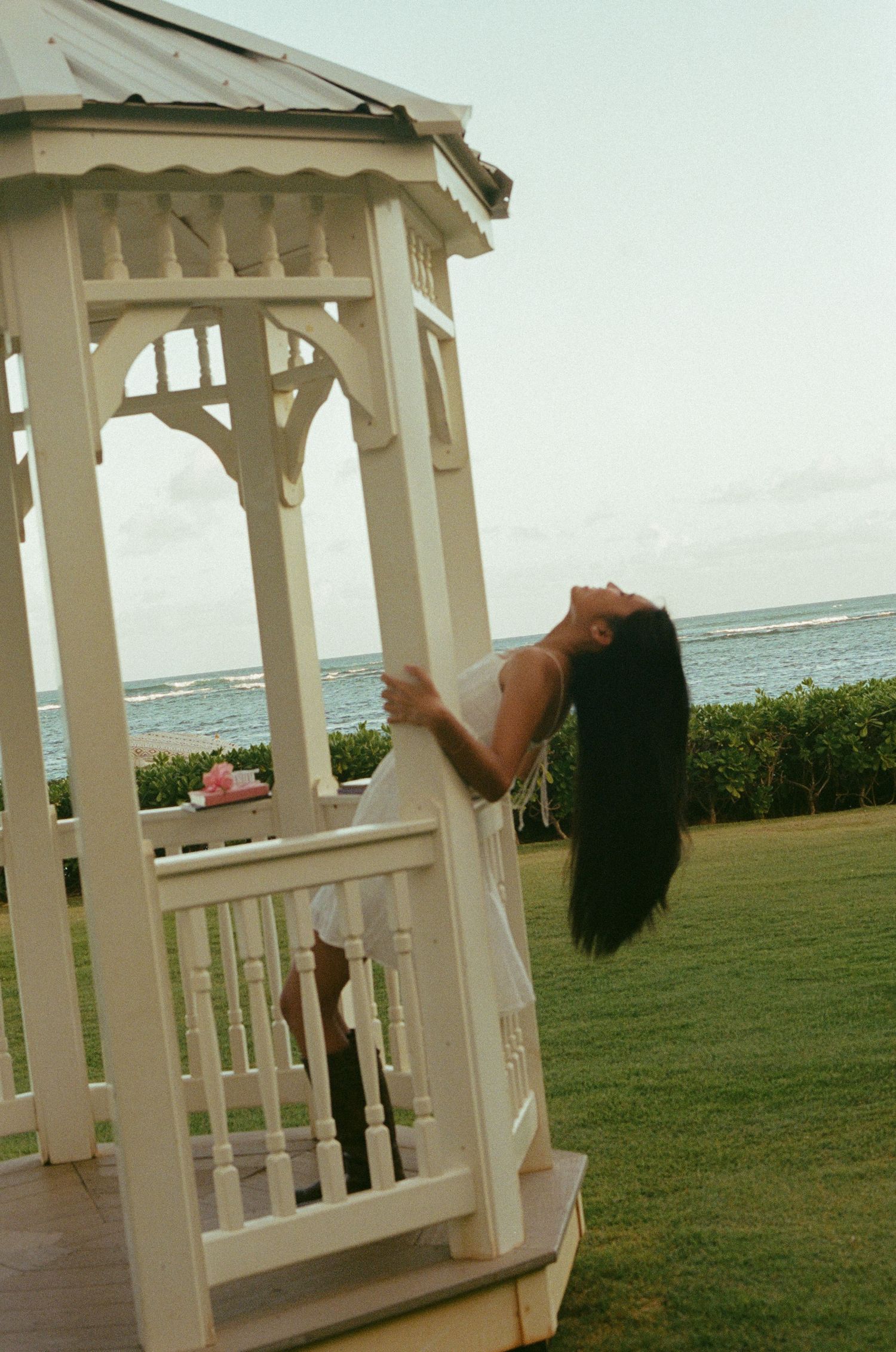 Woman in white dress leaning back gracefully in gazebo with ocean view in background.