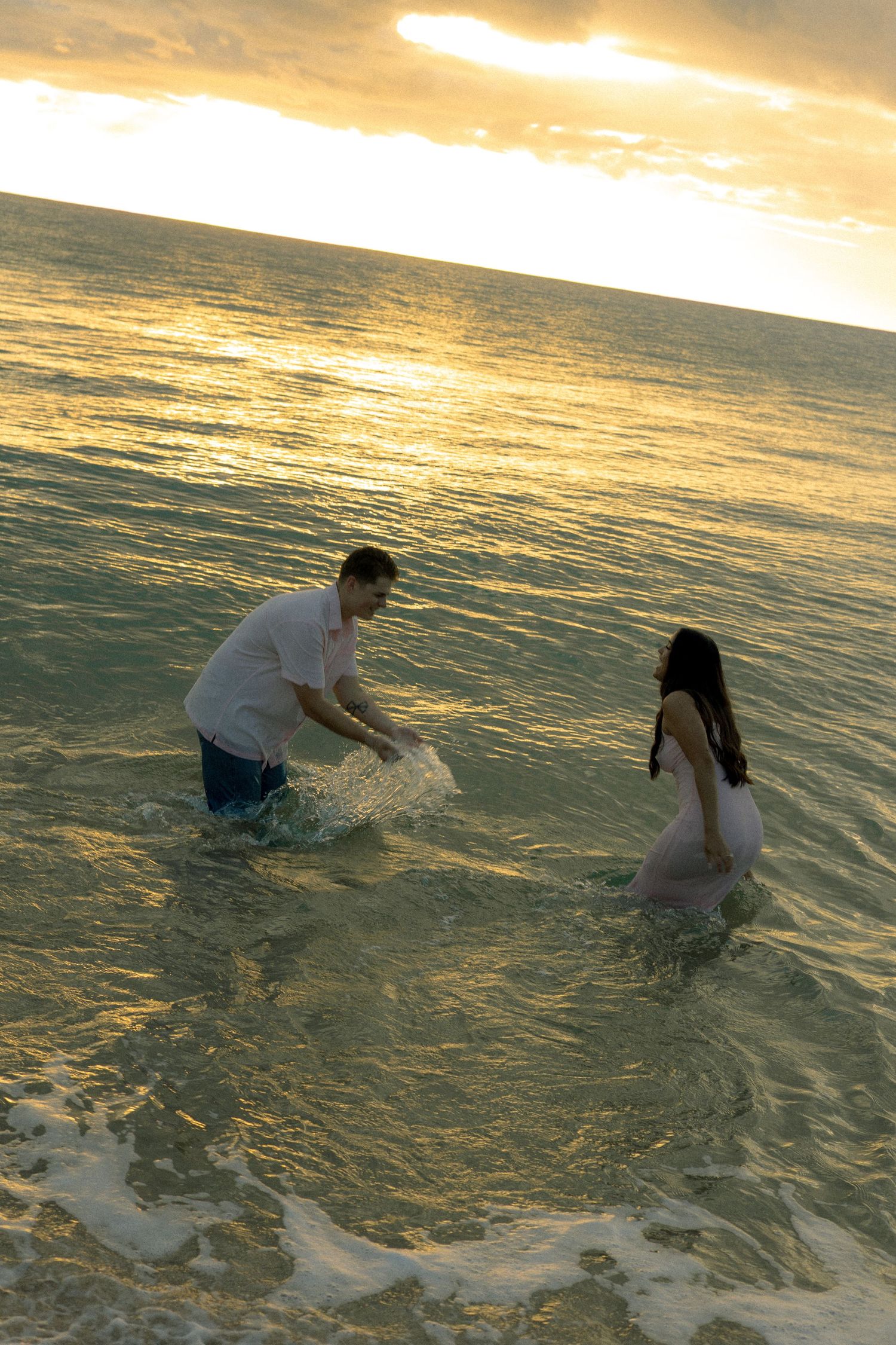 Couple playfully splashing in ocean waves during a golden sunset at the beach.