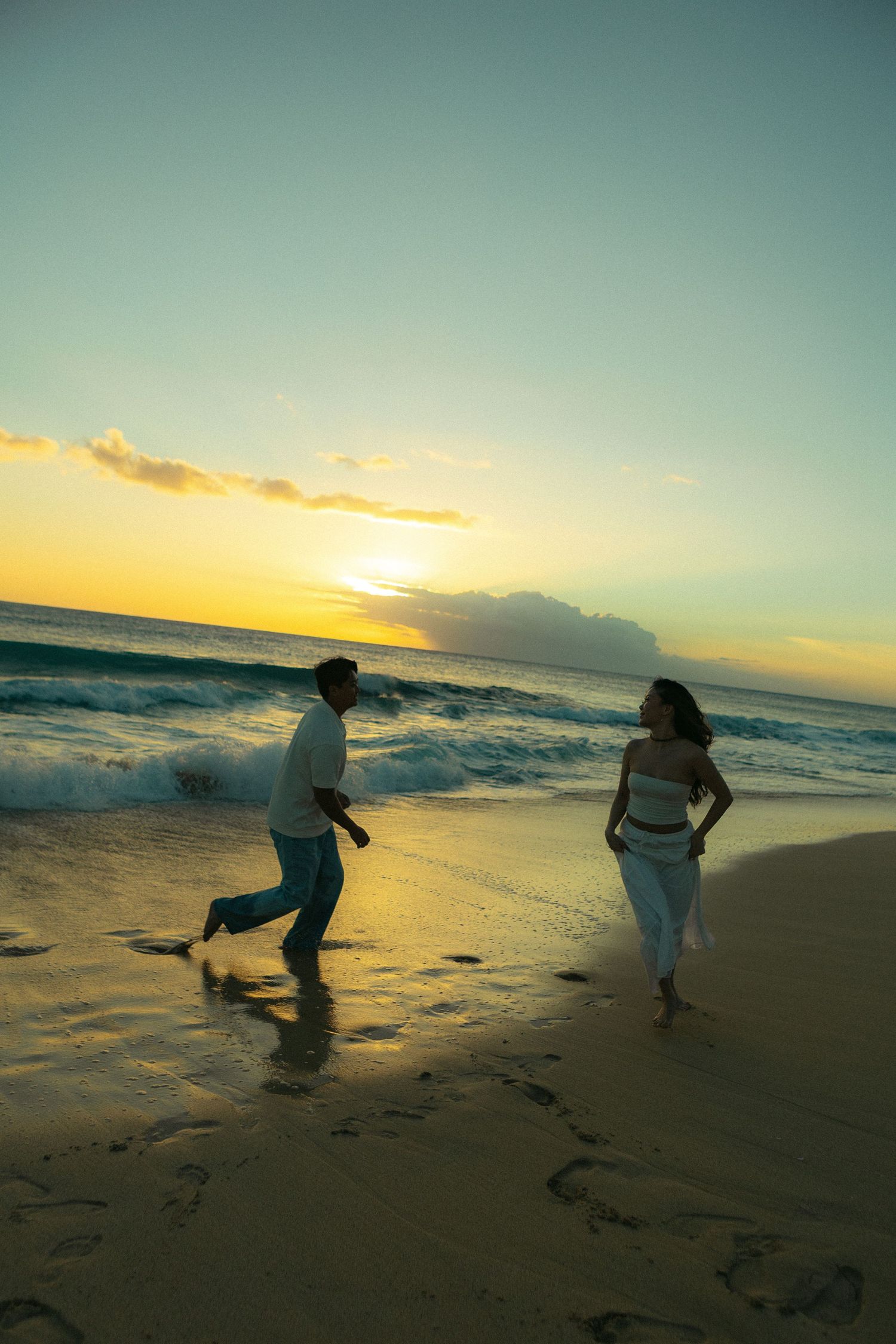 Couple running playfully on sandy beach at golden sunset with ocean waves in Hawaii.