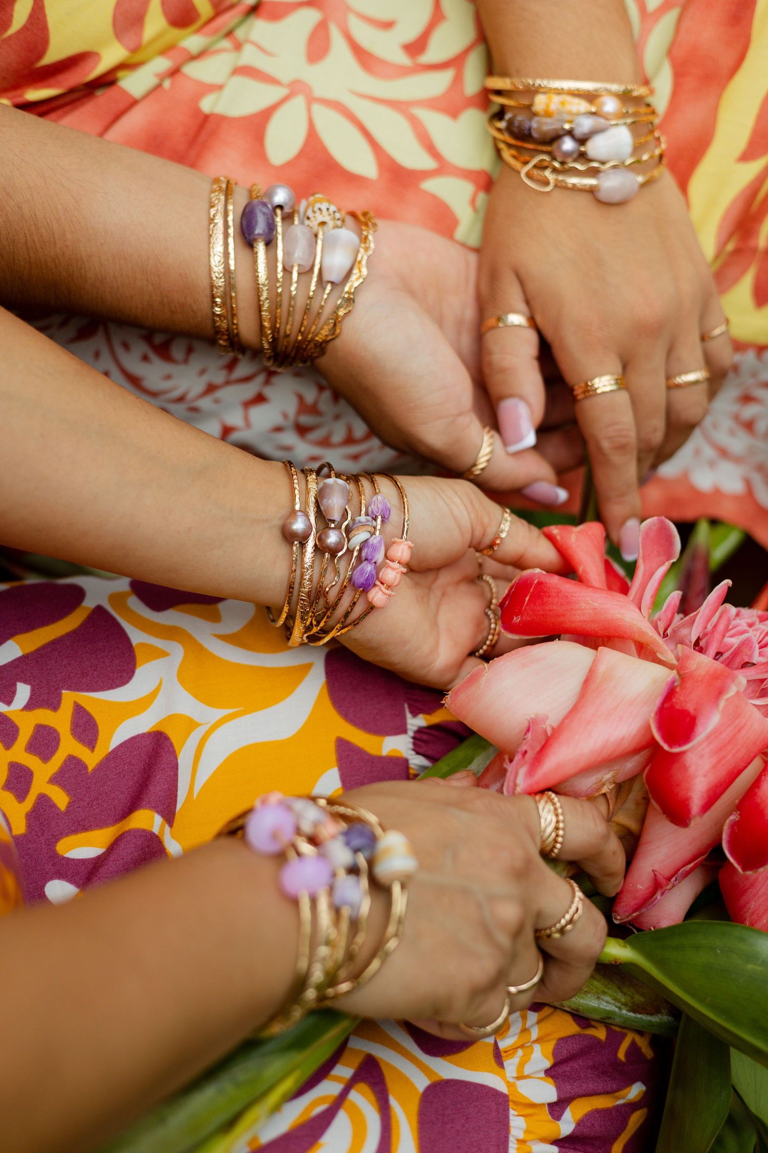 Multiple hands adorned with gold bangles and gemstone rings touching a pink tropical flower.