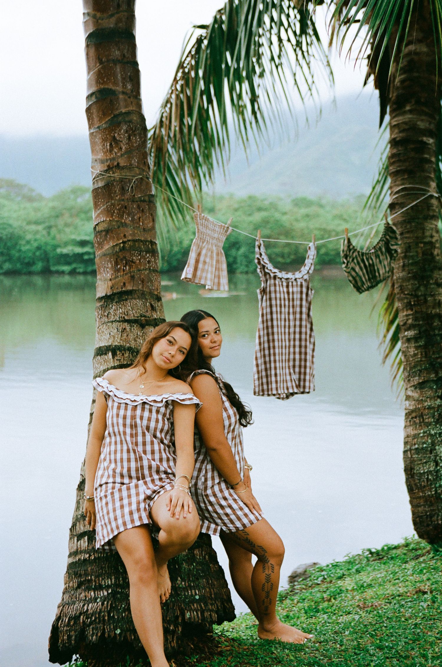 Two women in matching plaid dresses posing by palm trees near a serene lake.