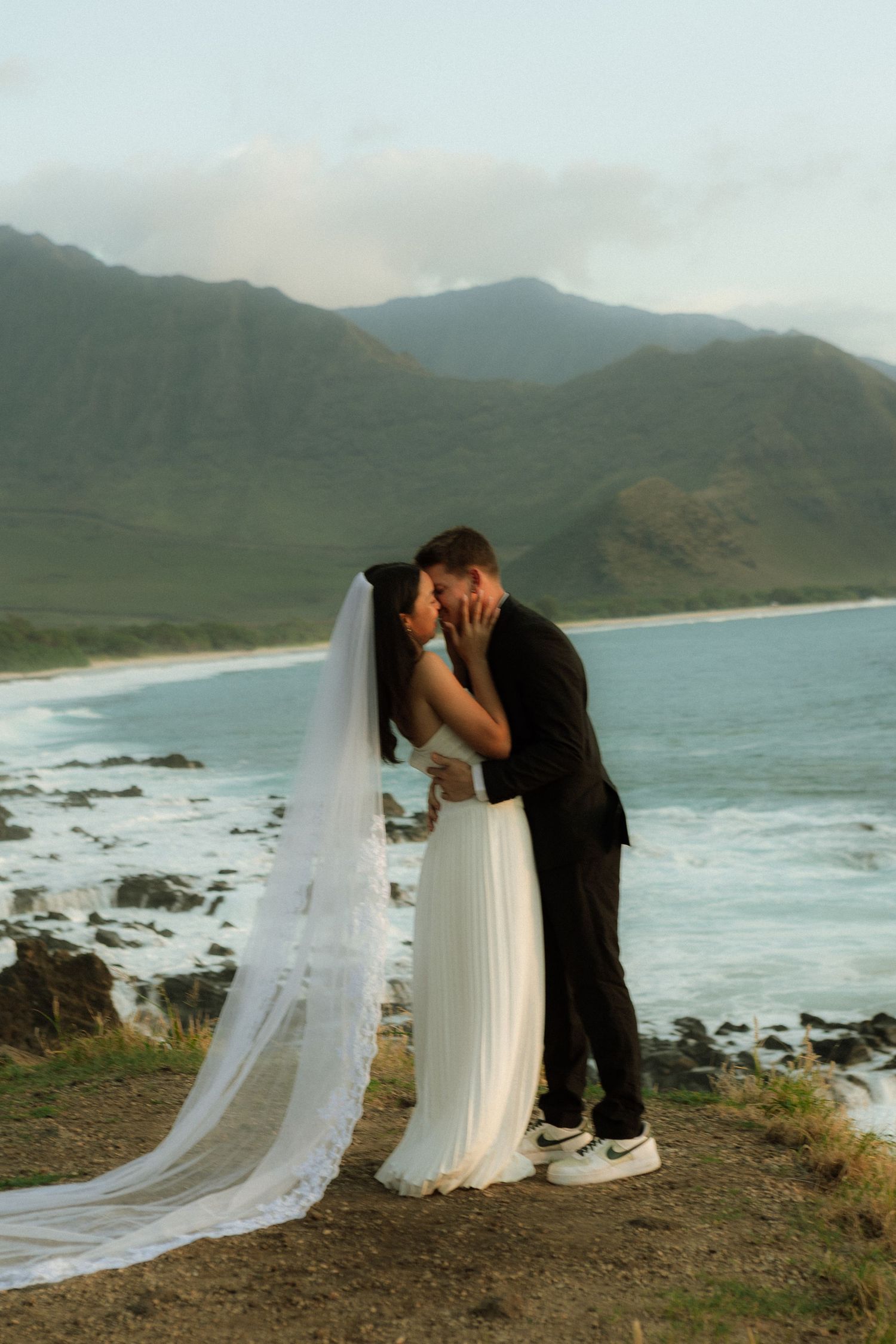 Bride and groom share a kiss on a coastal cliff in Hawaii, with turquoise ocean and green mountains behind them.