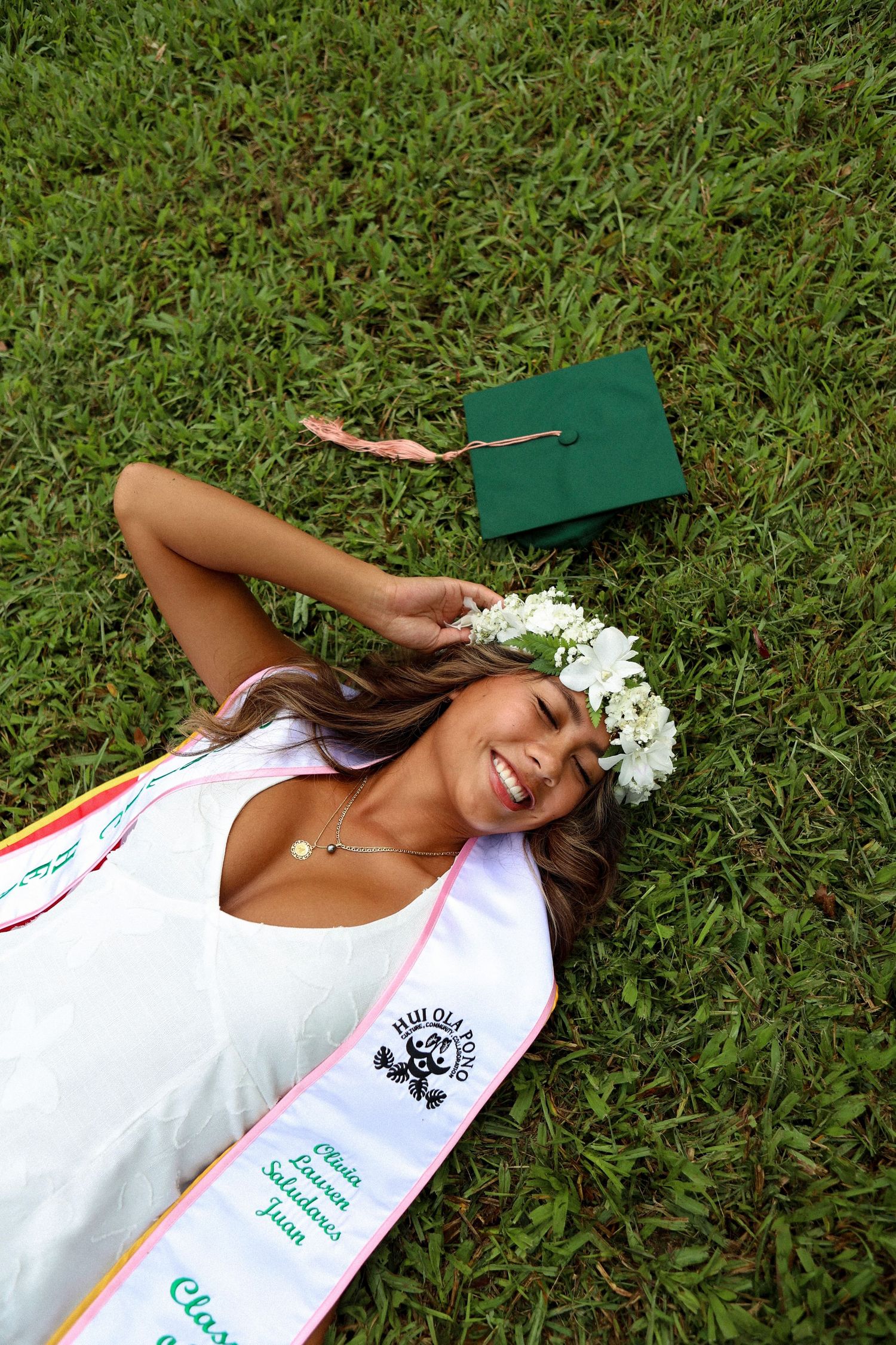 Graduate in white dress and flower crown laughs lying on grass with green cap.