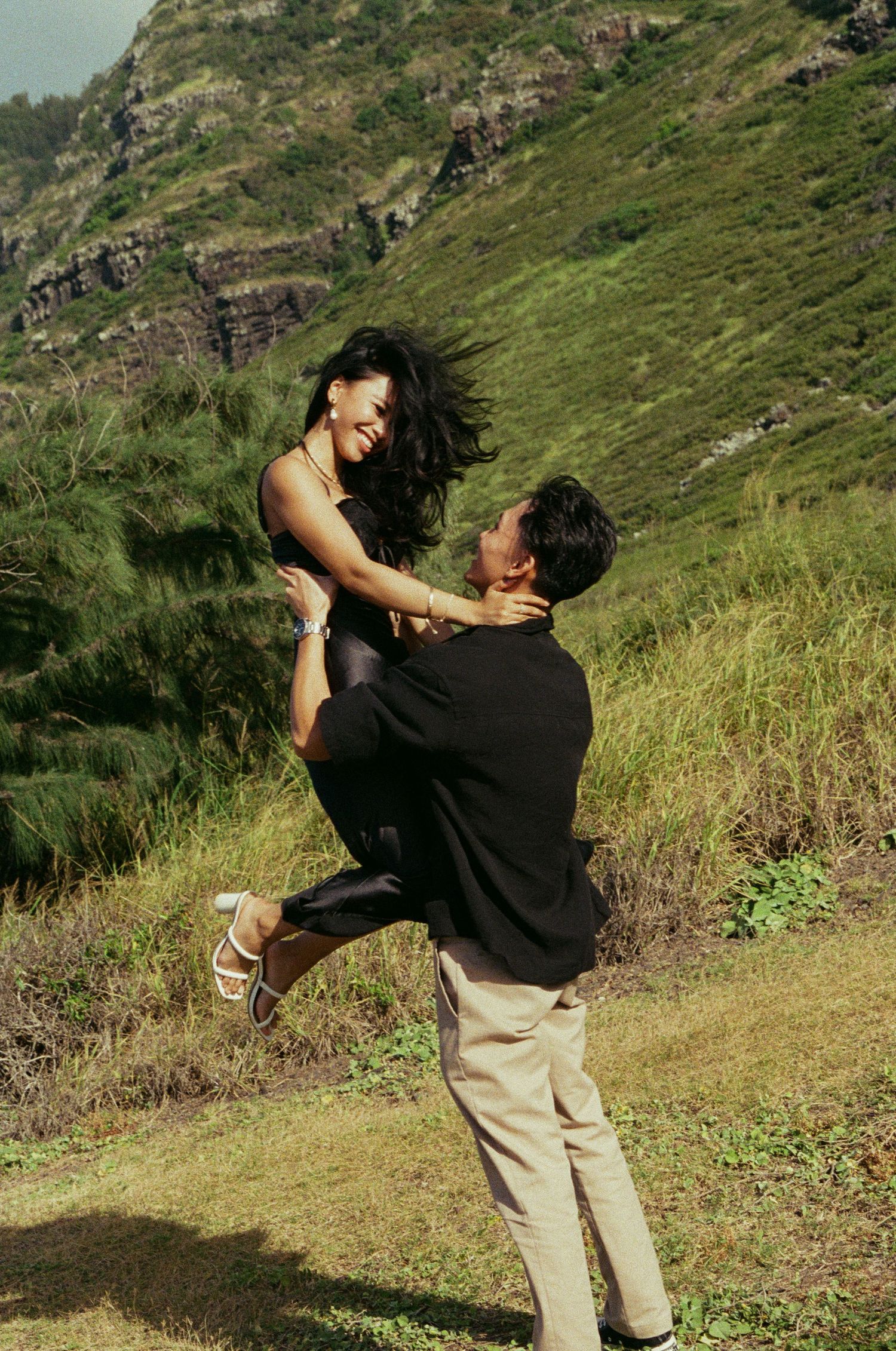 Man lifting and spinning smiling woman outdoors on a green hillside.