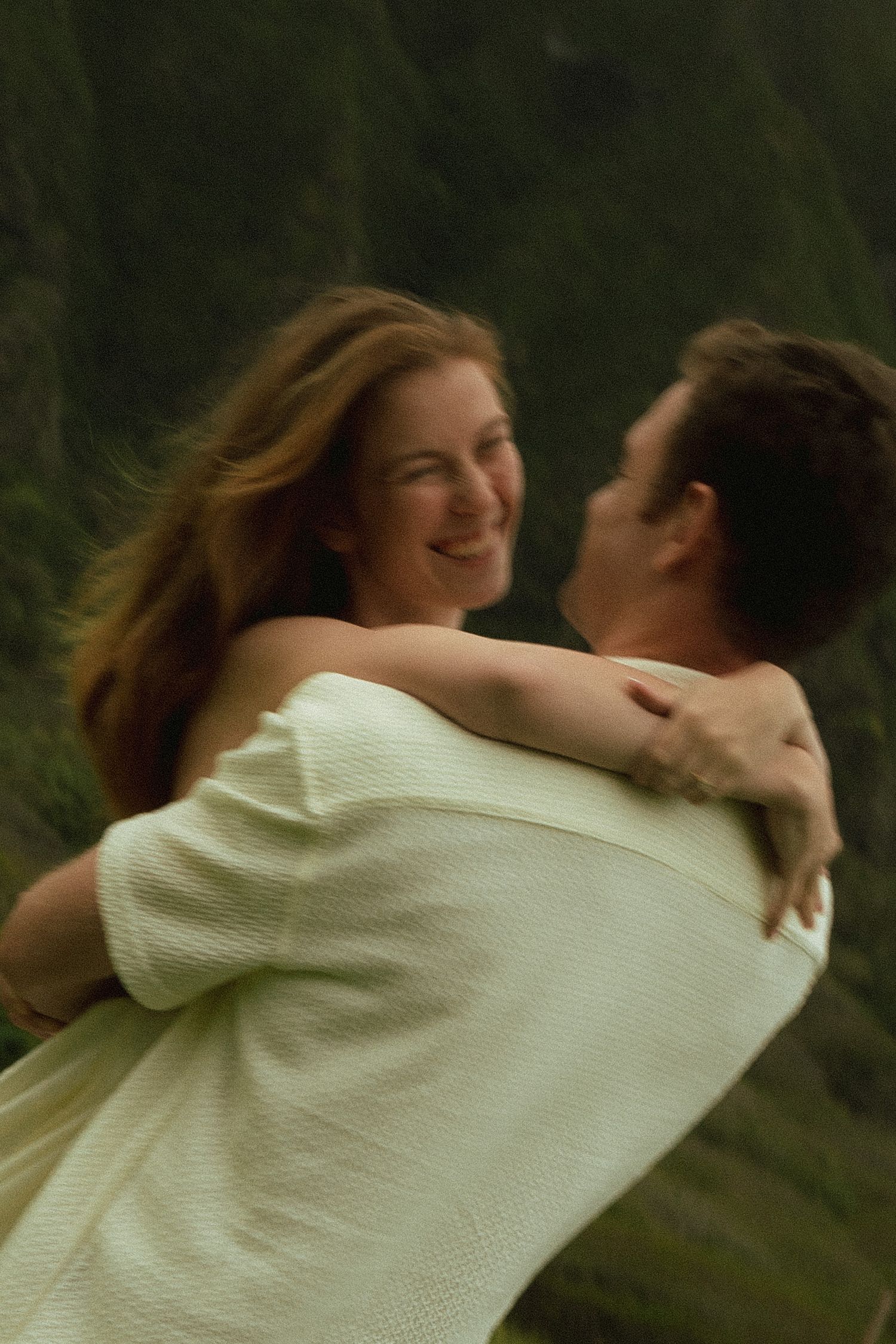 Man lifting smiling woman in white outfit against lush green mountain cliff background outdoors.