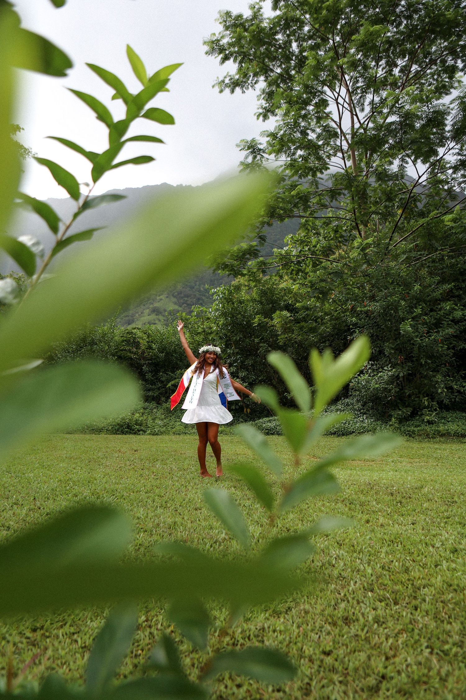 Woman in white dress celebrates in lush green Hawaiian valley with arms raised.