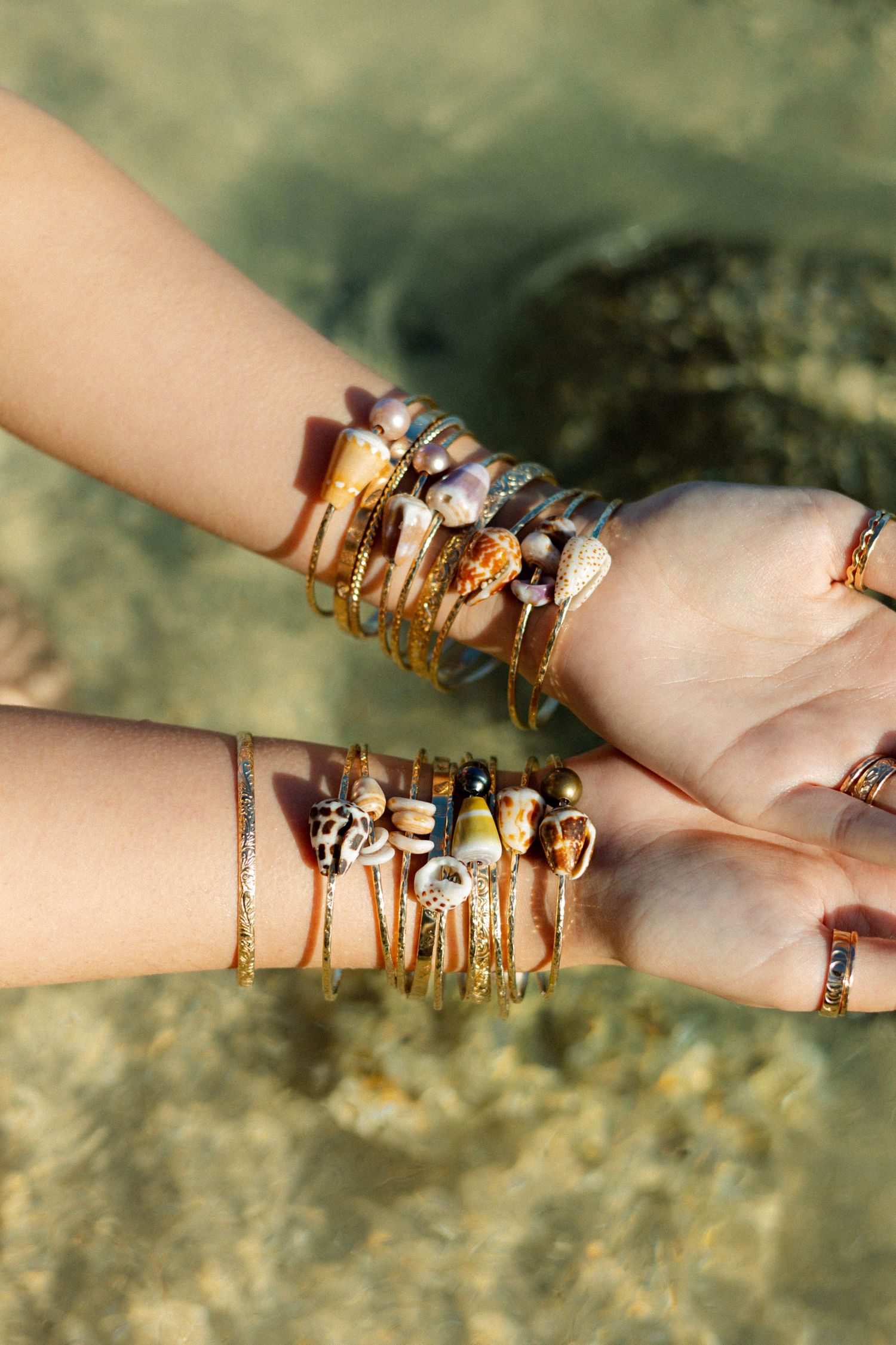 Two hands adorned with stacked gold and shell bracelets held over turquoise water.