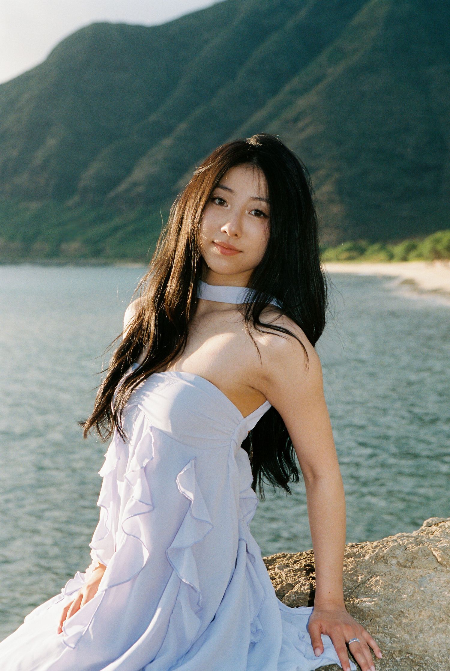 Young woman in white strapless dress sitting on rocks by a serene lake with green mountains in the background.