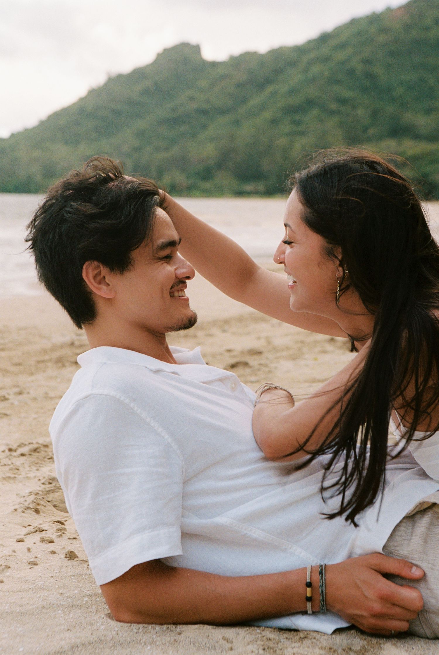 Couple sitting on sandy beach smiling at each other, lush green mountains and calm water in background.