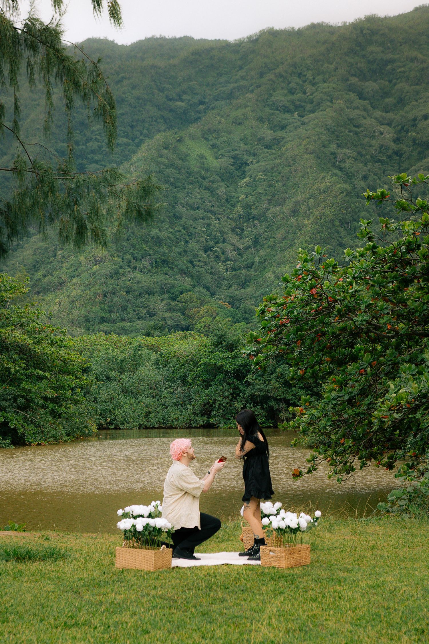 Man kneeling to propose to woman outdoors near pond with white flowers and lush green mountains backdrop.