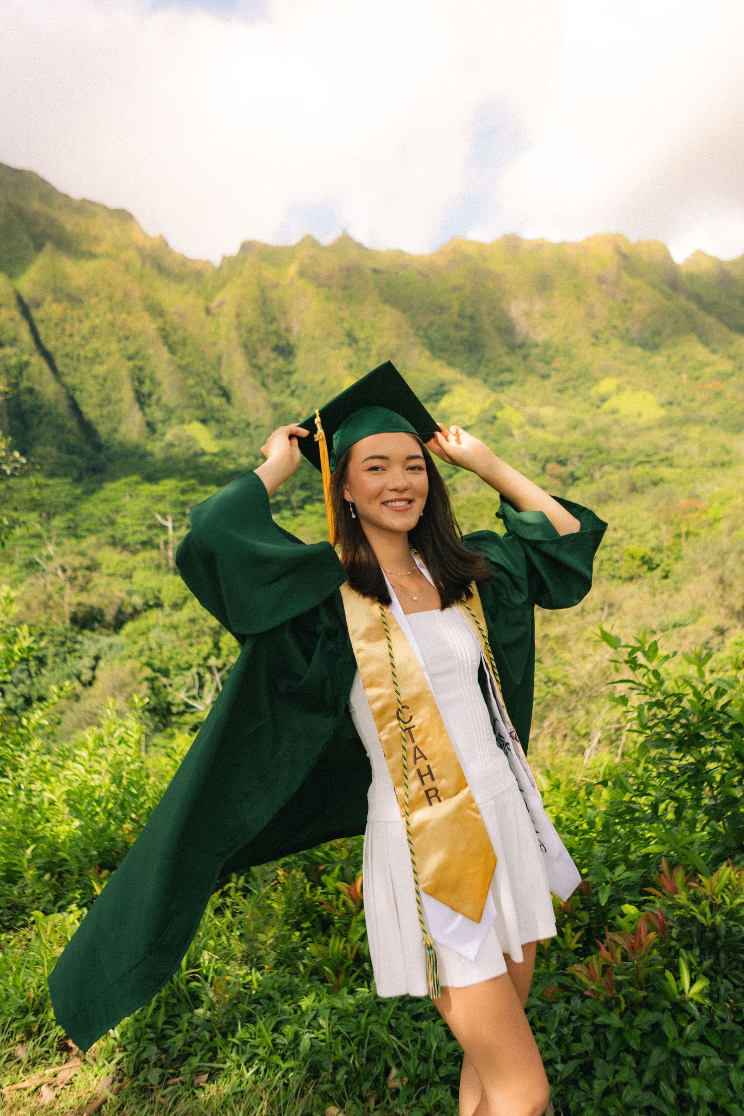 Smiling graduate in green cap and gown with gold sash posing before lush mountain landscape.
