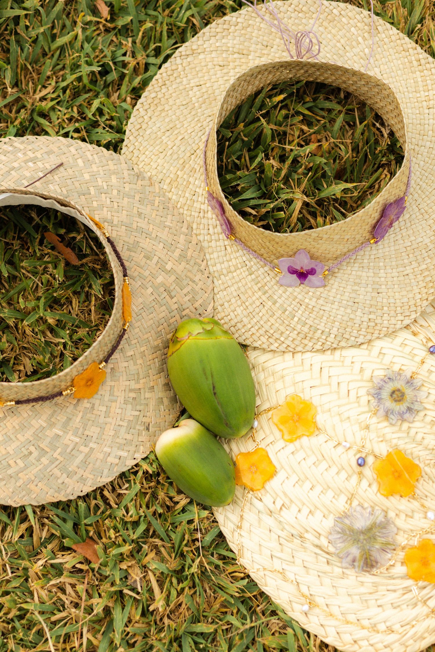 Three straw hats with flower decorations laid on green grass with tropical fruits.