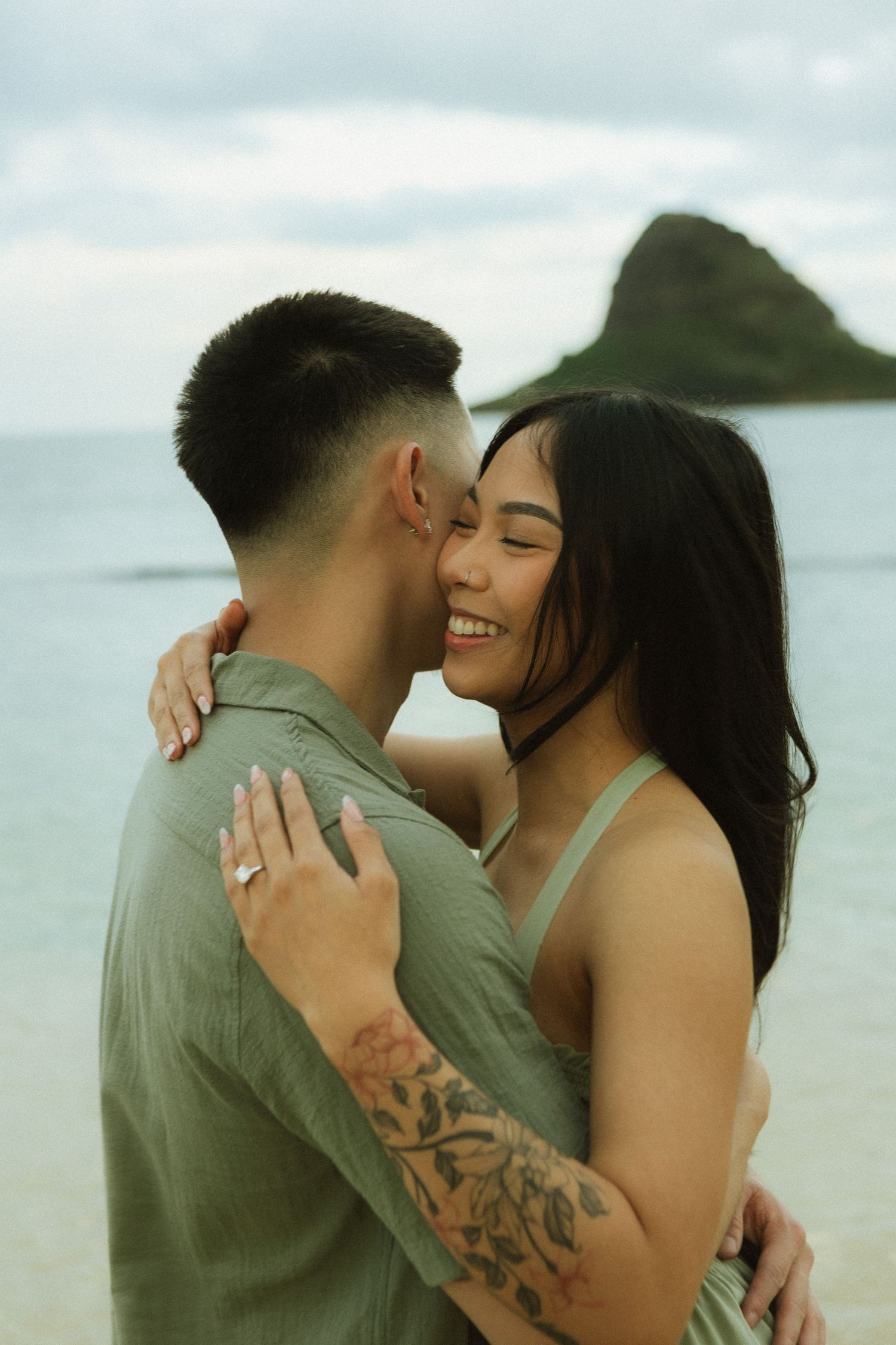 Smiling woman with engagement ring embracing her partner on a cloudy beach in Hawaii.
