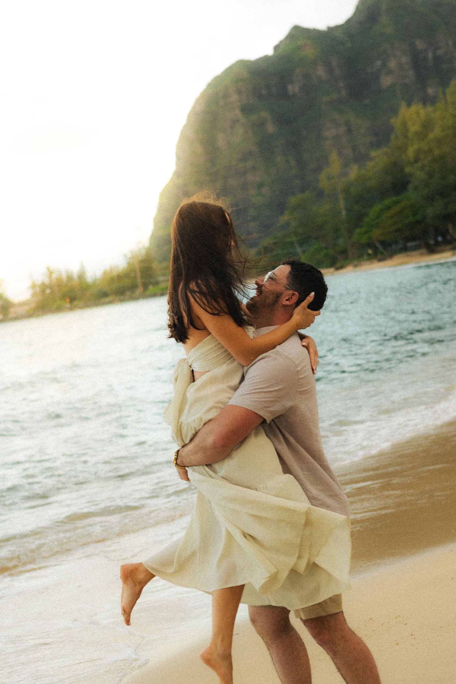Couple embracing on a tropical beach at sunset with lush green mountains in the background.
