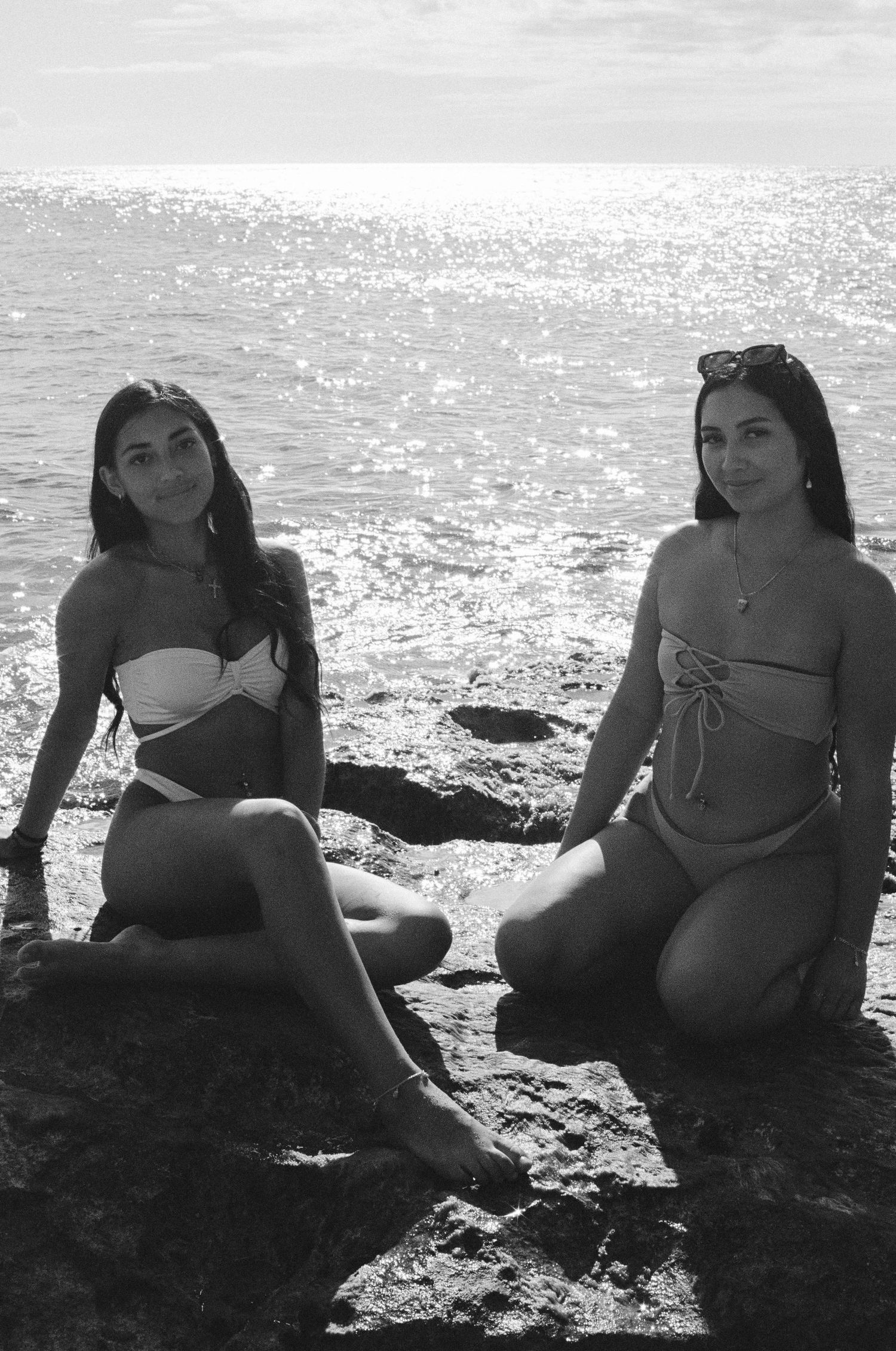 Black and white photo of two women in bikinis sitting on rocks by sparkling ocean.