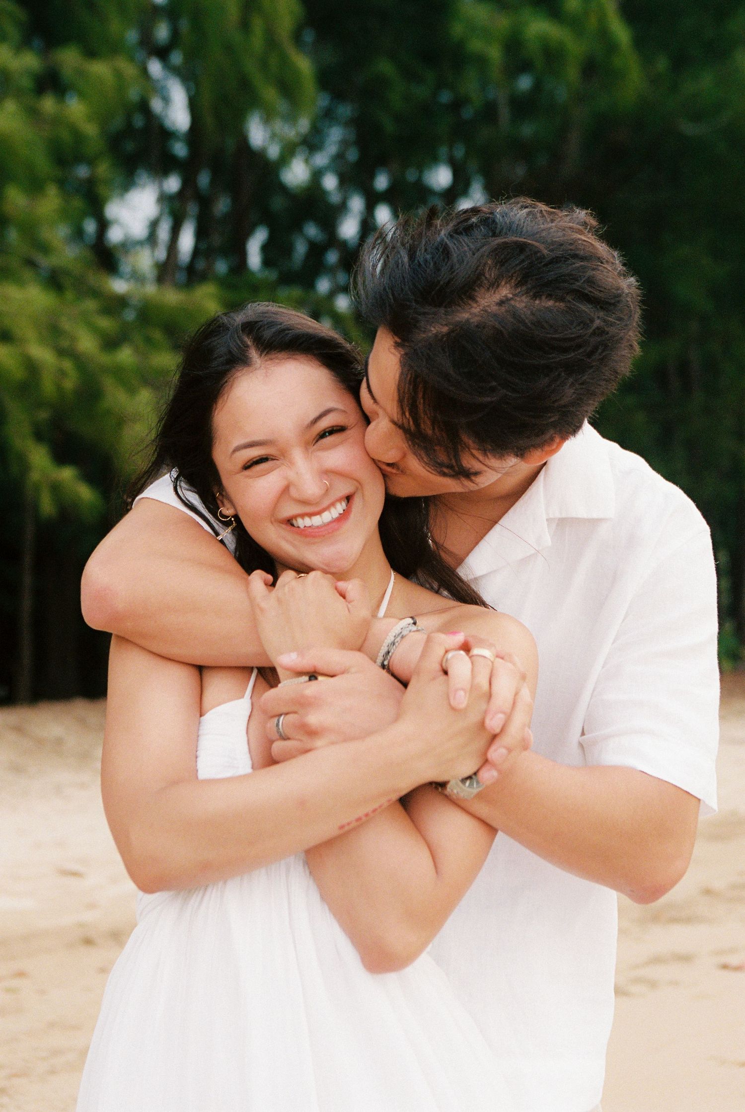 A smiling woman in a white dress is embraced and kissed on the cheek by a man on a sandy beach.