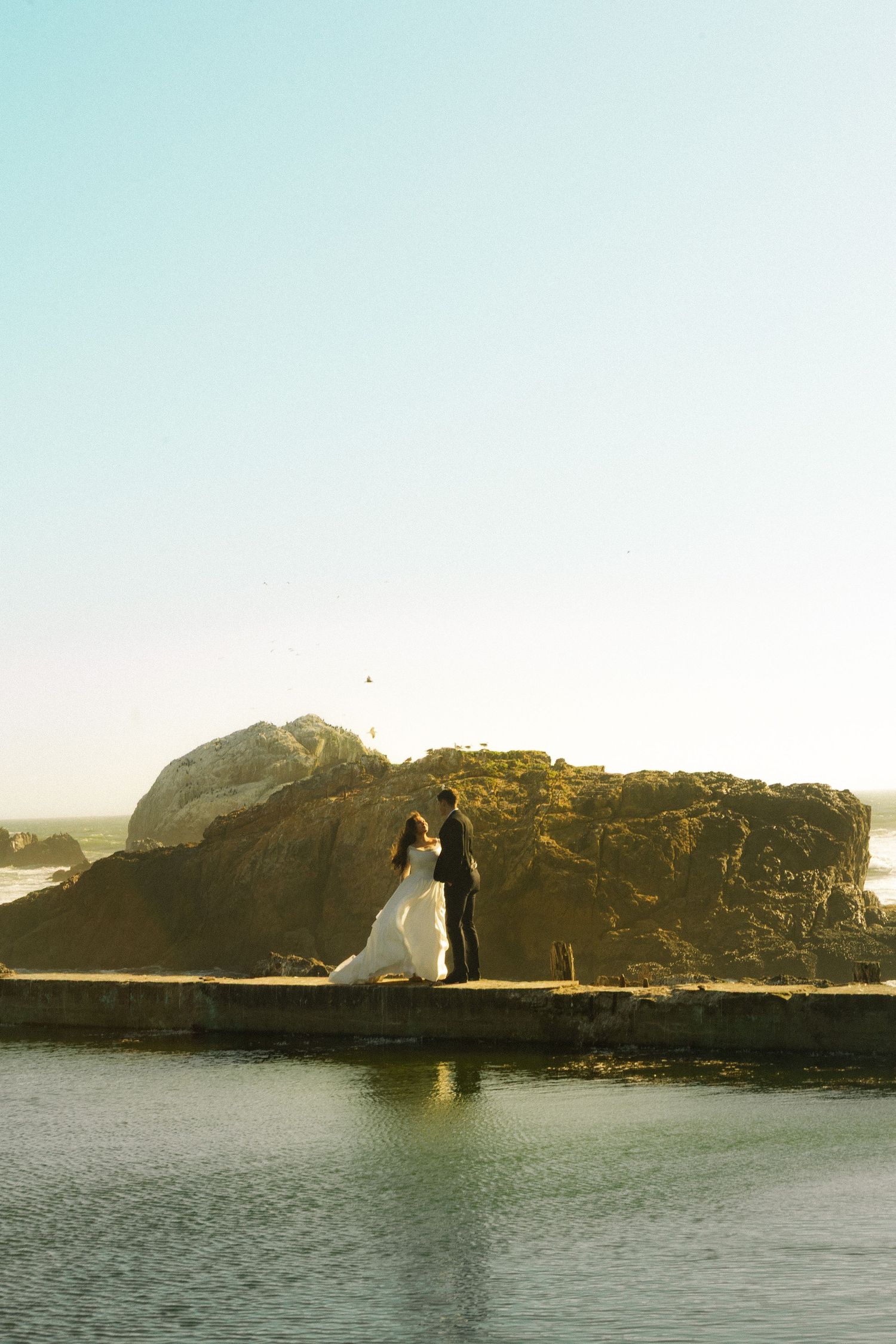 Bride and groom standing on a coastal rock ledge by the ocean during golden hour at their beach wedding.