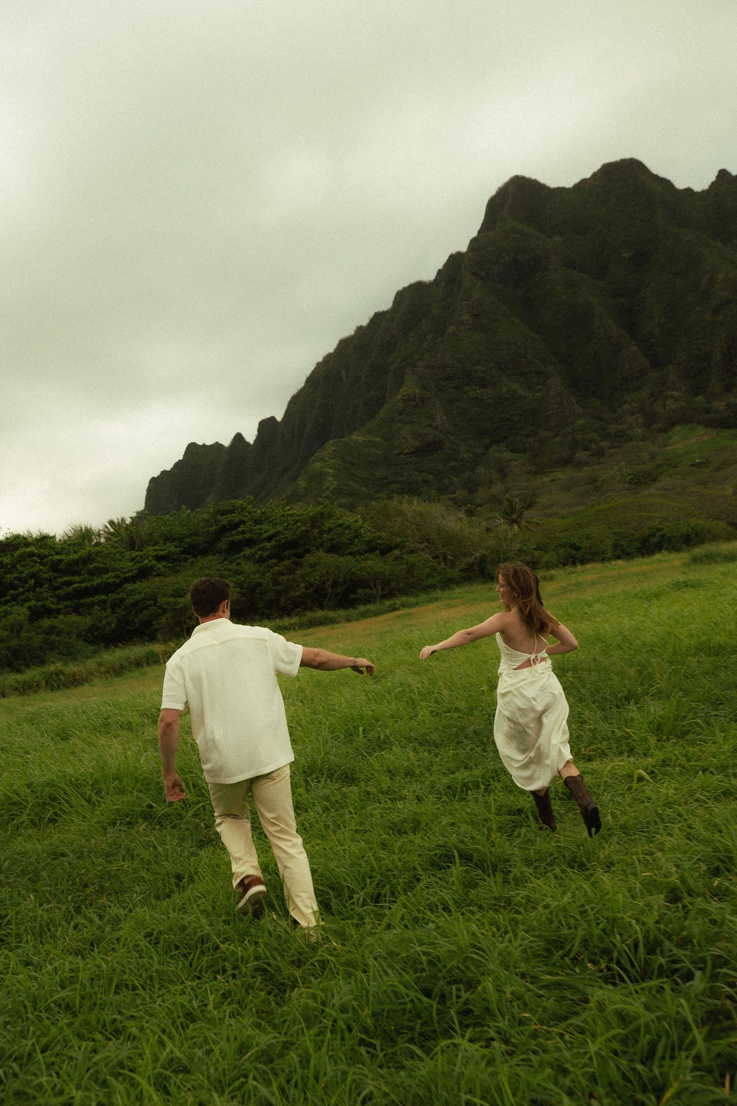 Couple holding hands running through green meadow with dramatic rocky mountain peaks in background.