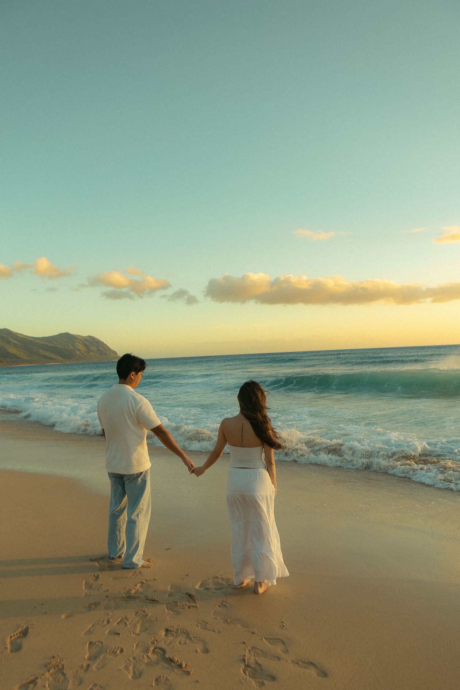 Couple holding hands on a sandy beach at sunset, waves gently rolling in with mountains visible on the horizon.