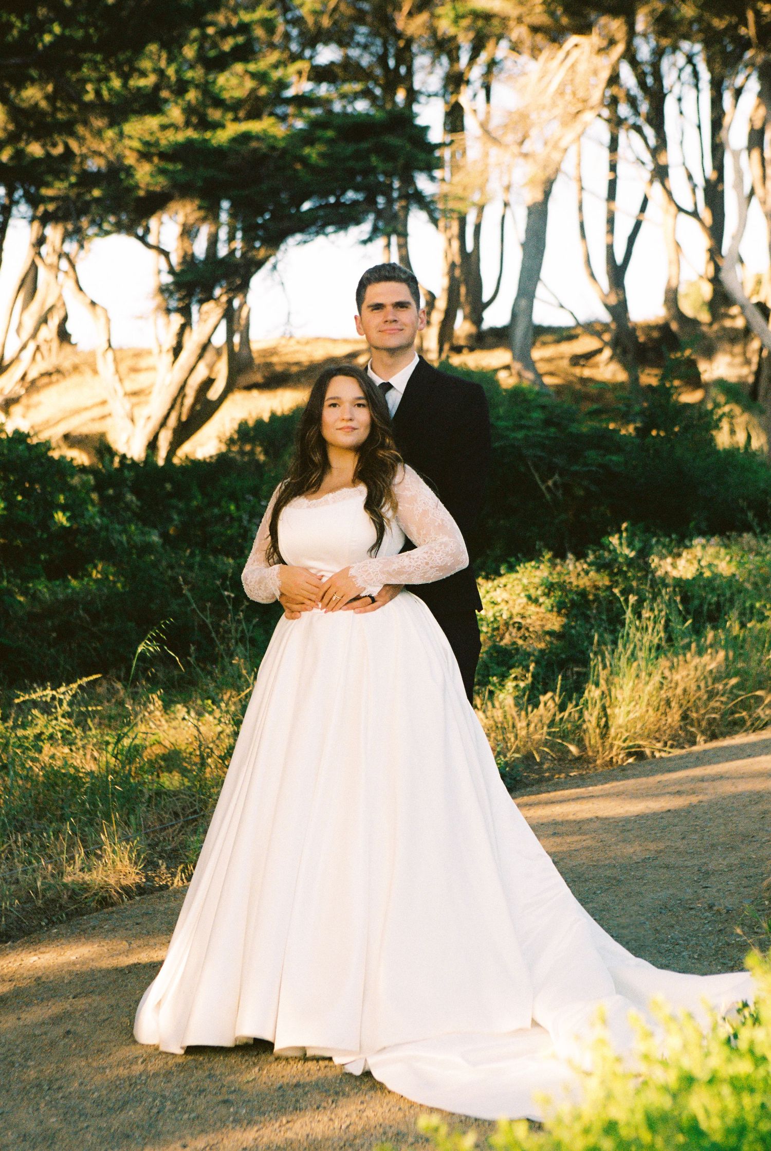 Bride in white long-sleeve gown and groom in black suit pose together outdoors among trees at golden hour.