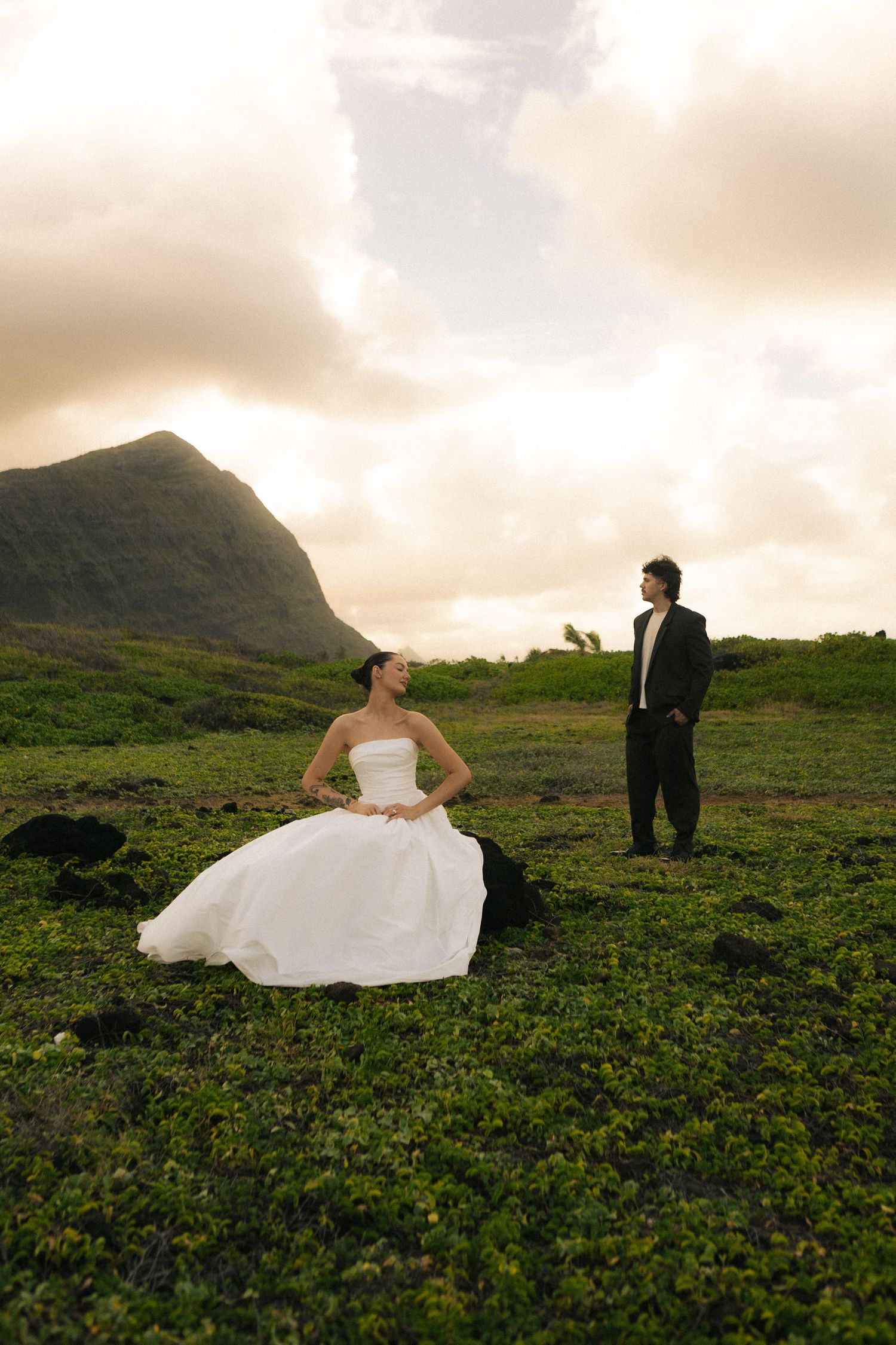 Bride in white gown sits on green field while groom in black tuxedo stands nearby with mountain backdrop.