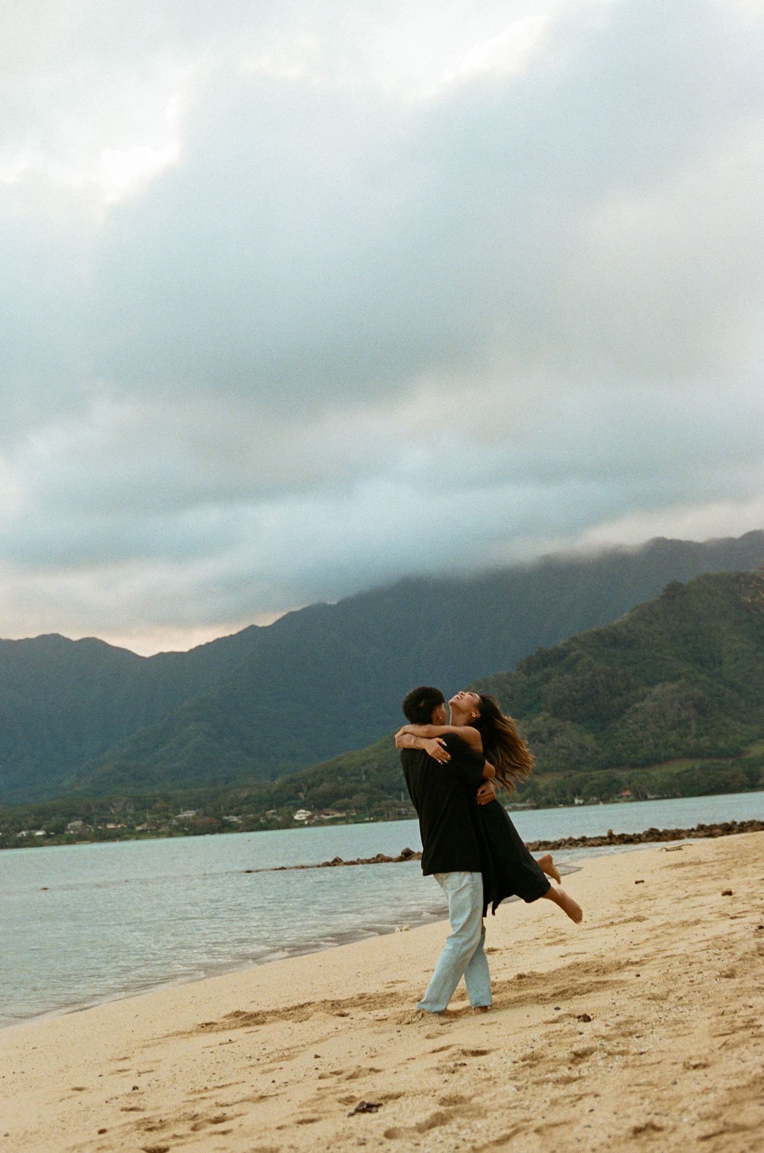 Couple embracing joyfully on Hawaiian beach with mountains and dramatic cloudy sky behind them.