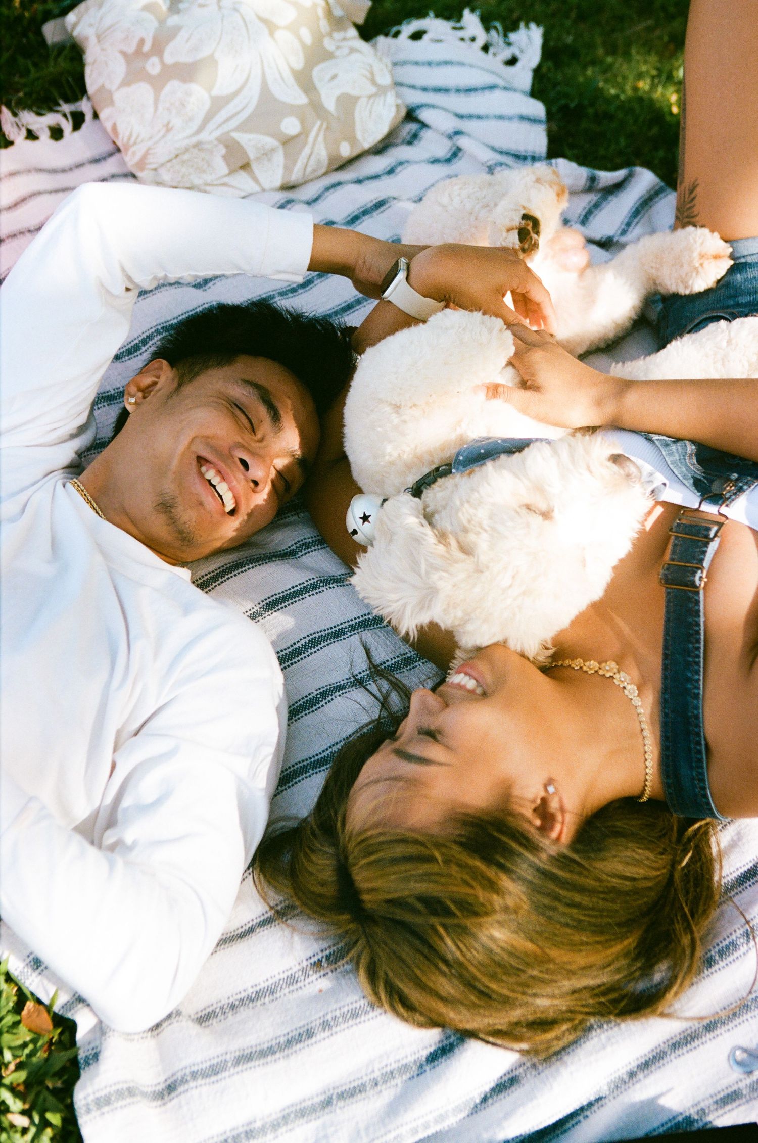 Smiling couple lying on blanket outdoors with fluffy white dog, enjoying sunny day together.