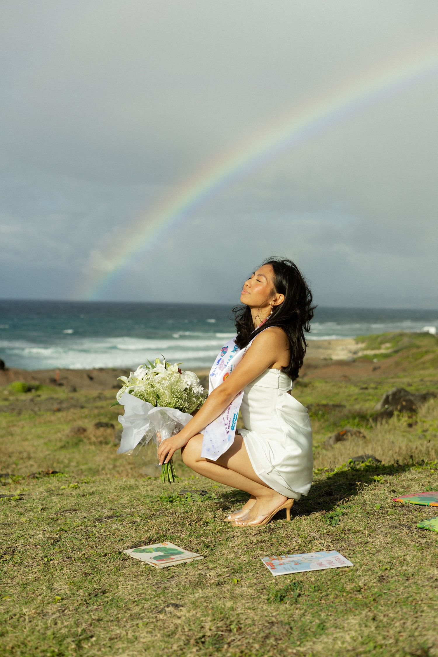 Woman in white dress holds bouquet kneeling by ocean with rainbow in background in Hawaii.