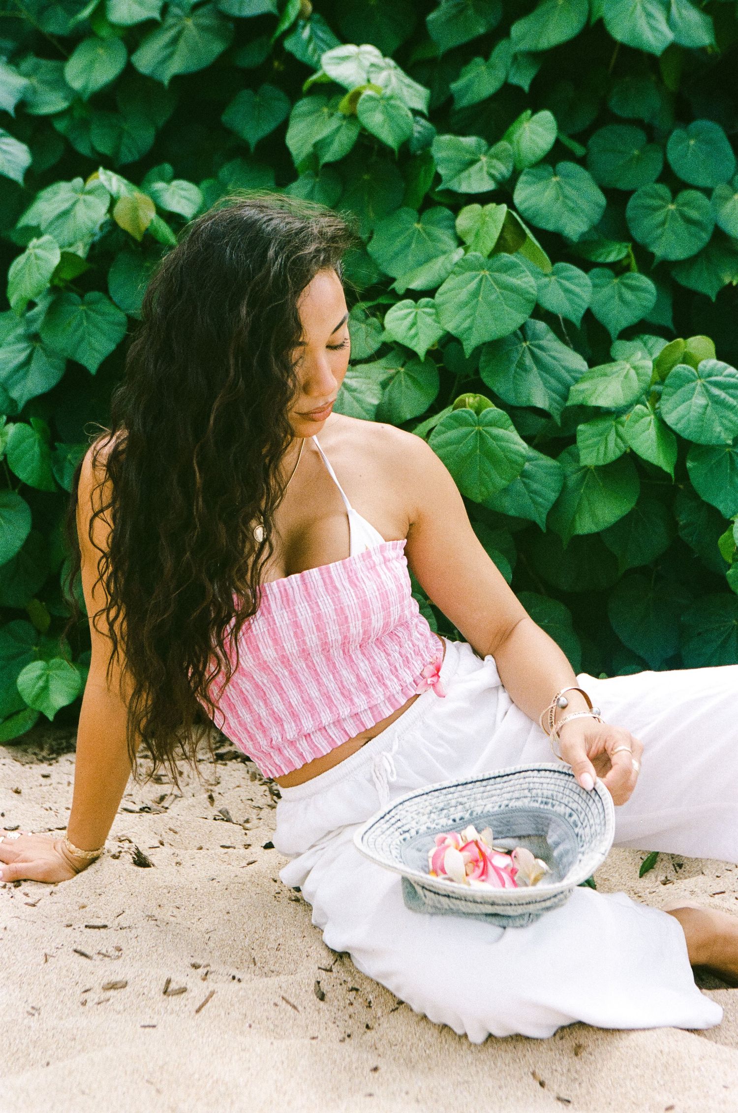 Woman in pink striped crop top and white pants sitting on sand against lush tropical green foliage.