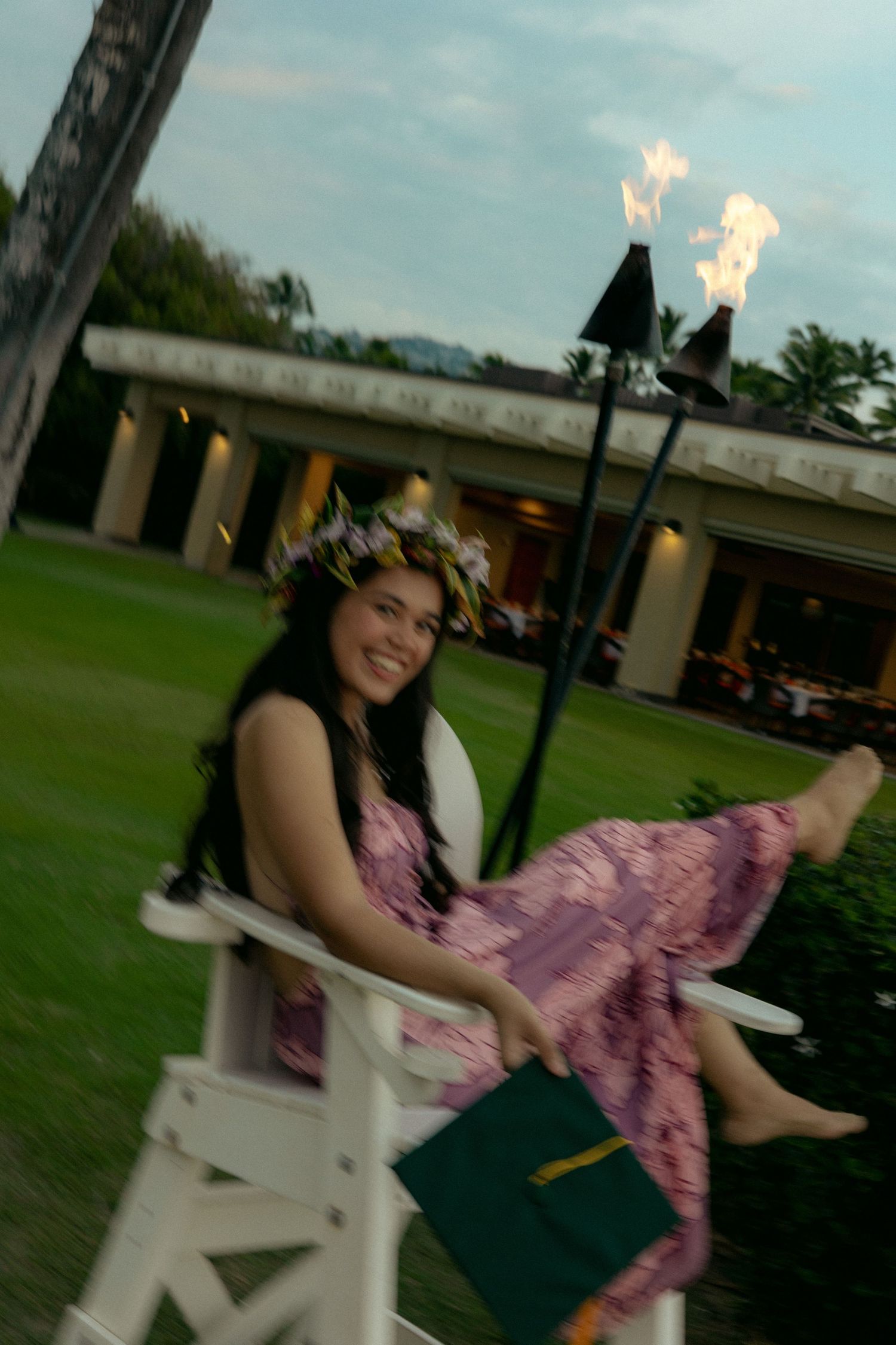 Smiling woman in floral crown sits in white chair near tiki torches at dusk.