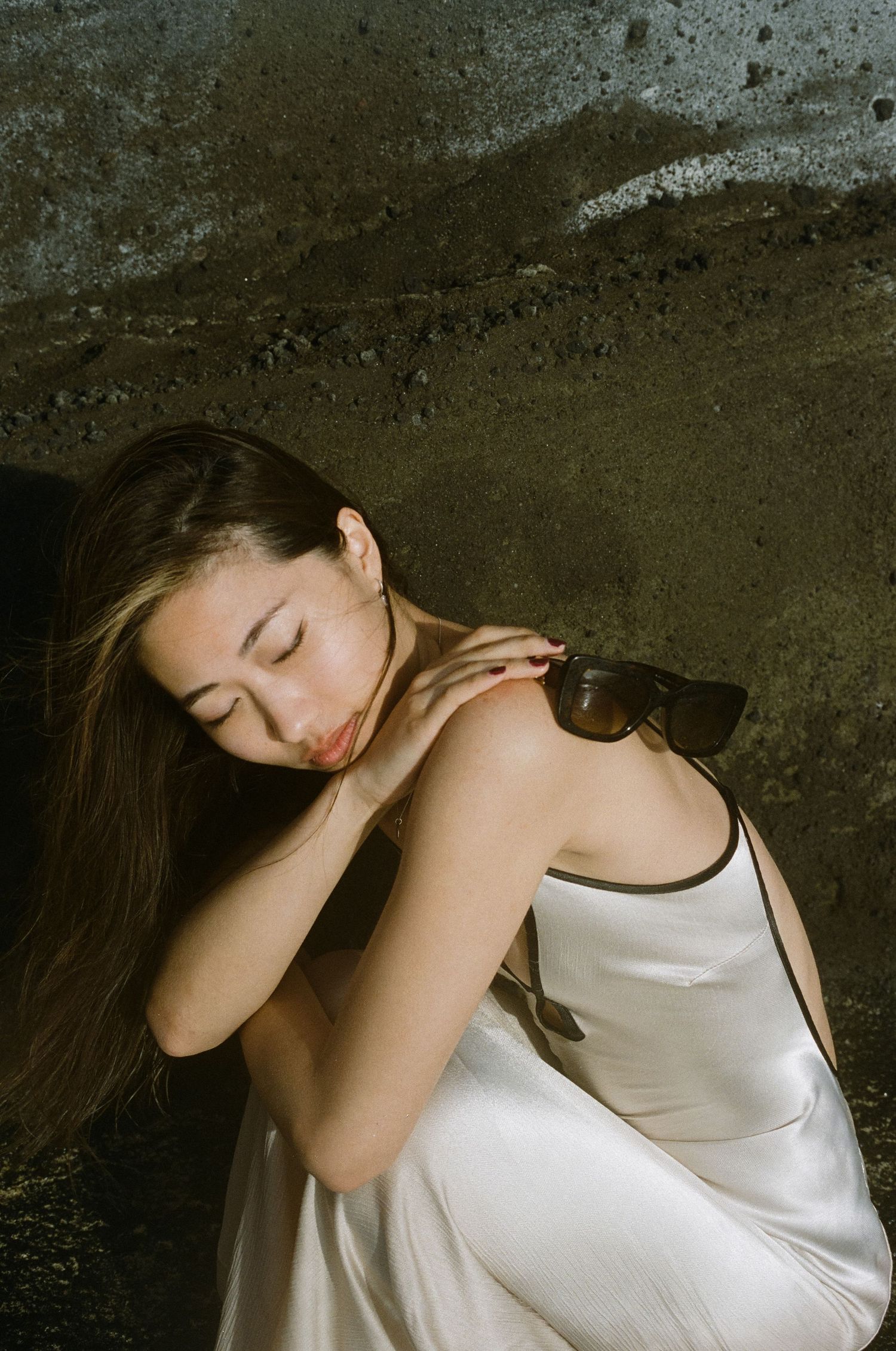 Asian woman in white satin slip dress crouching against dark wall holding sunglasses.