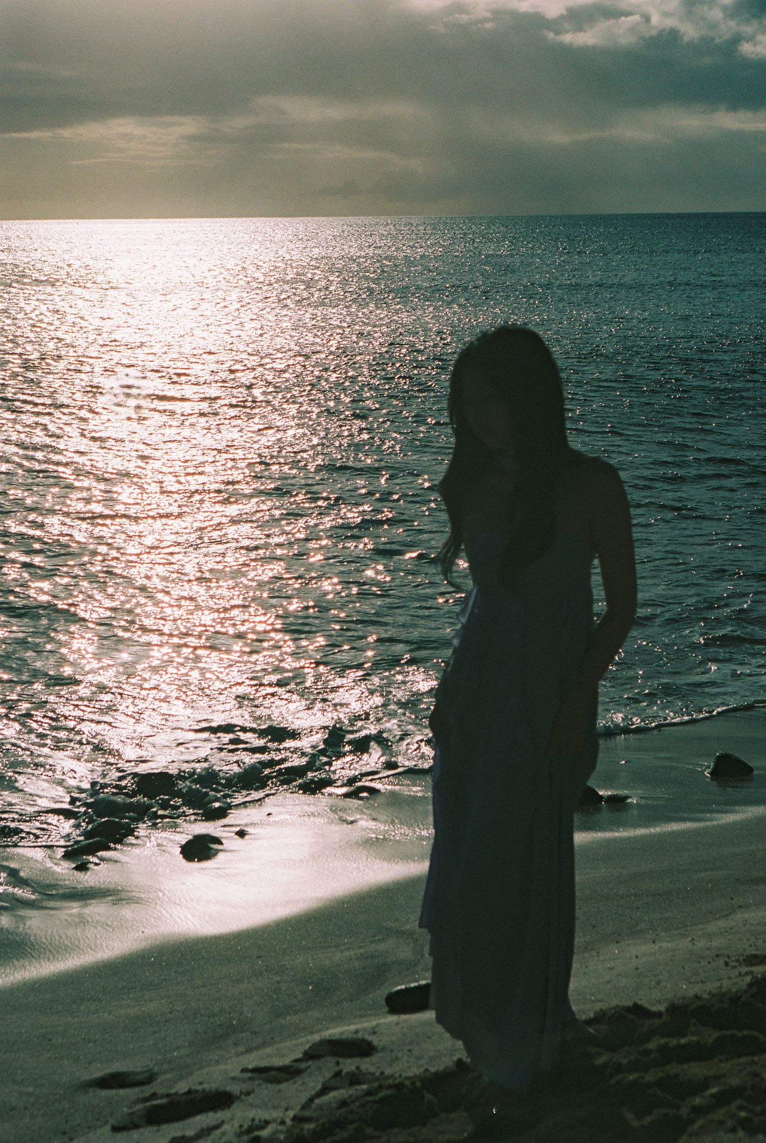Silhouette of a woman in a long dress standing on a beach at sunset with shimmering ocean reflections.