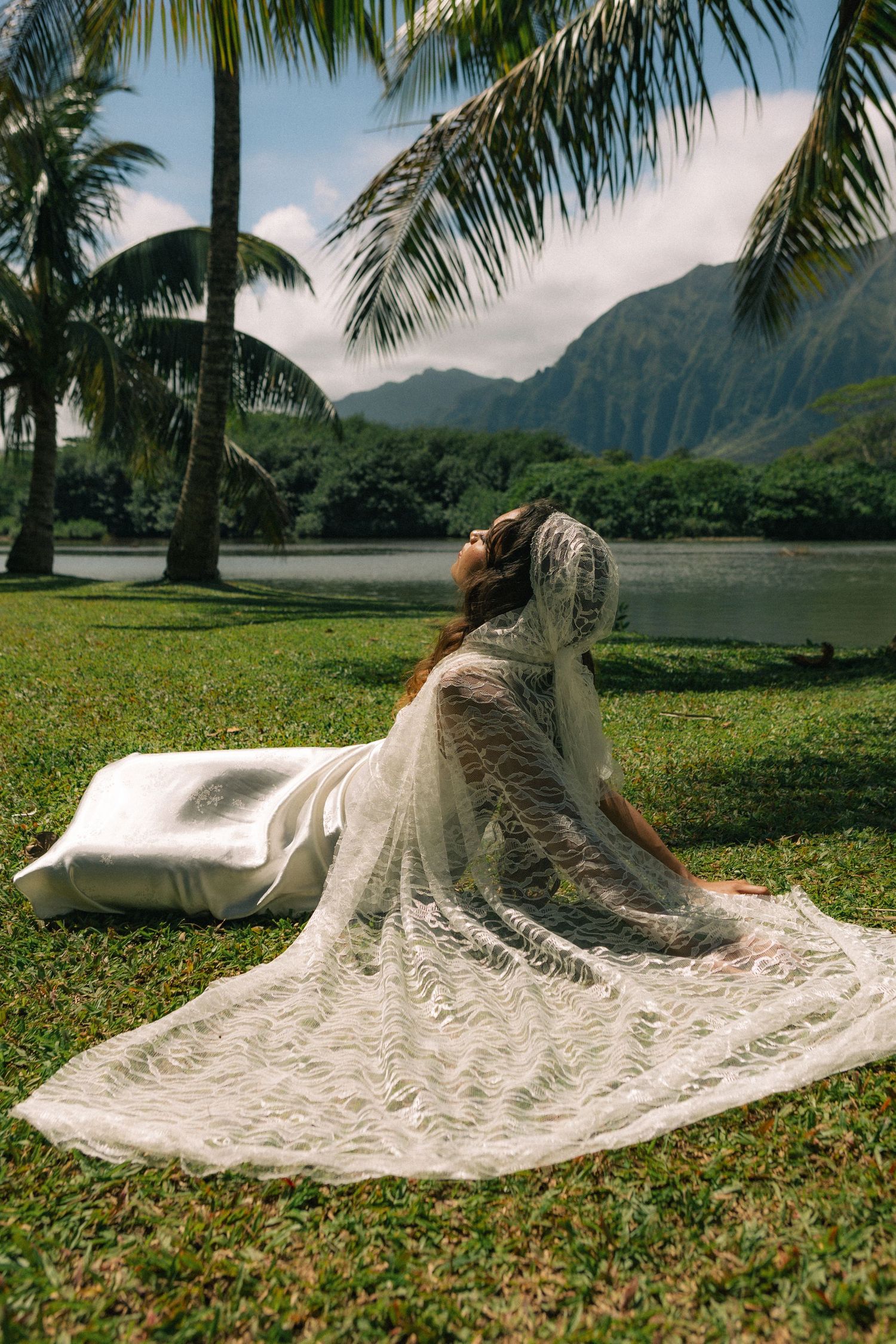 Bride in lace wedding gown sitting on grass near tropical lake with palm trees and mountains in Hawaii.