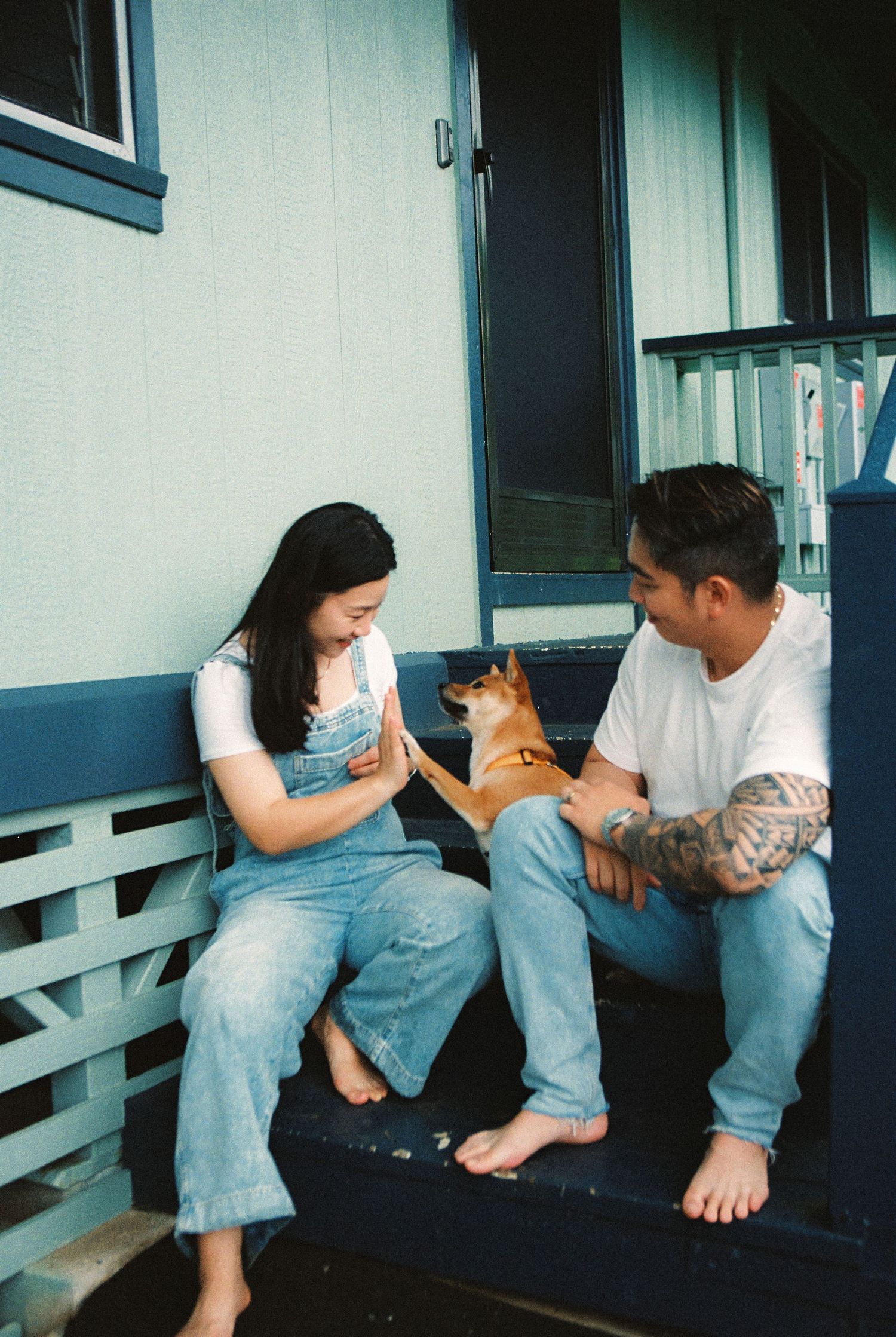 Couple sitting on porch steps with a Shiba Inu dog, woman in denim overalls, man with tattoos, casual outdoor setting.