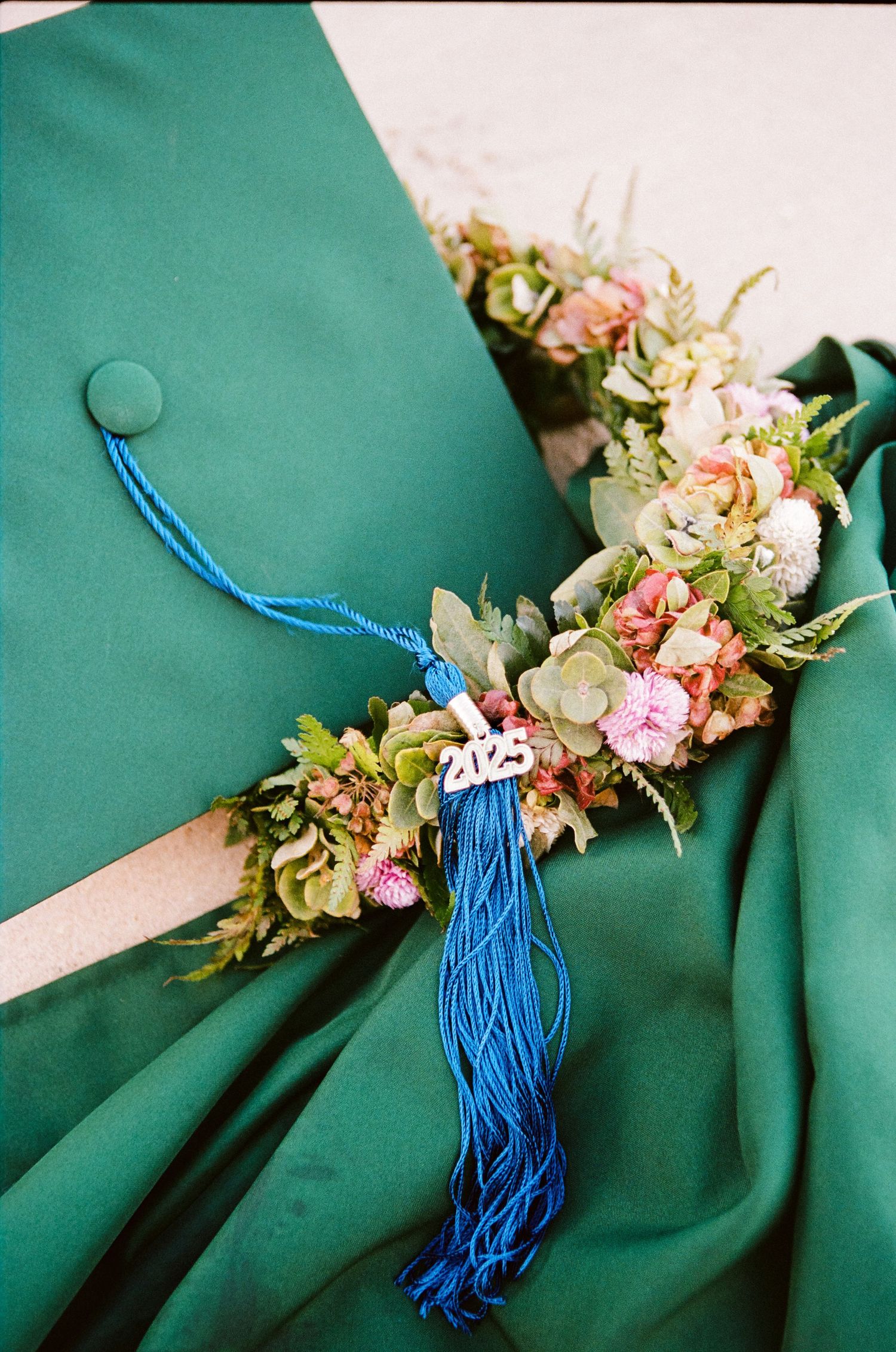 Green graduation cap with blue tassel and floral crown decorated with pink and white flowers.