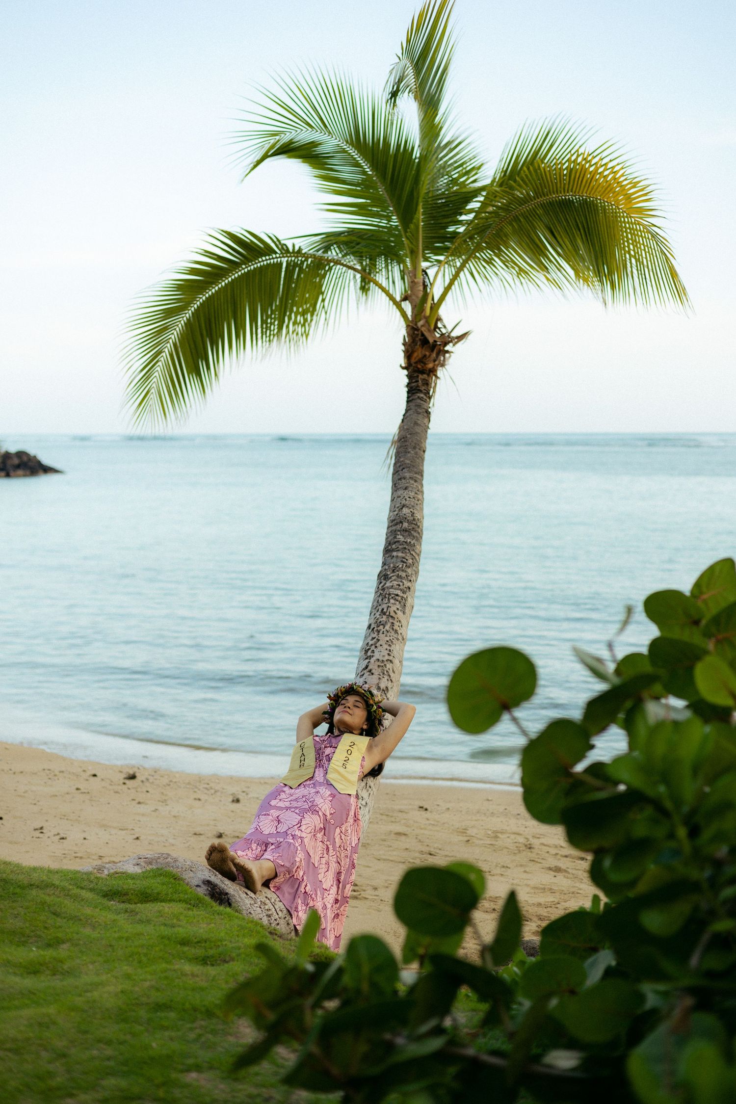 Woman in pink floral dress leaning against a leaning palm tree on a tropical Hawaiian beach.