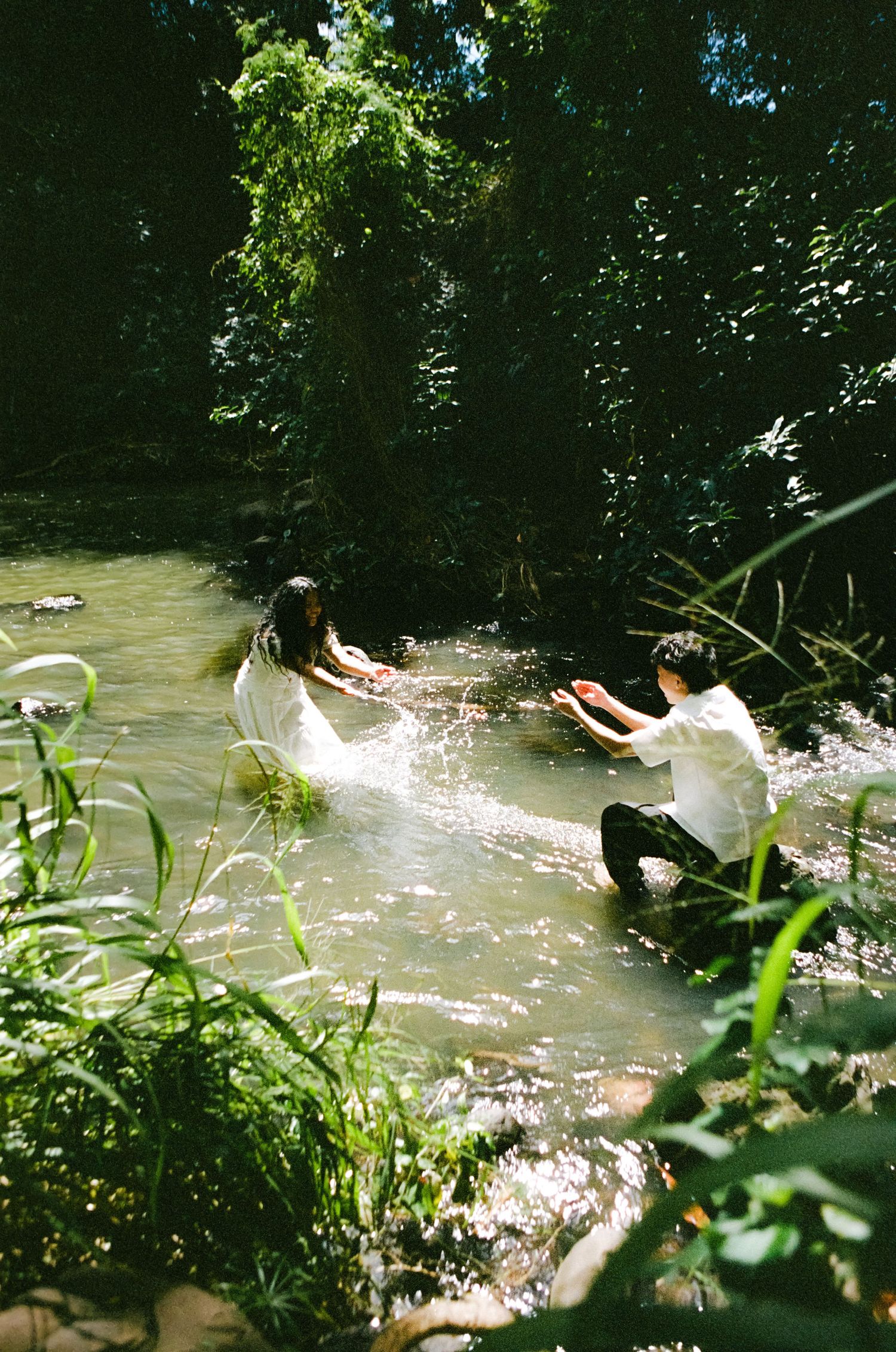 Couple playfully splashes in a sunlit forest stream surrounded by lush green trees.