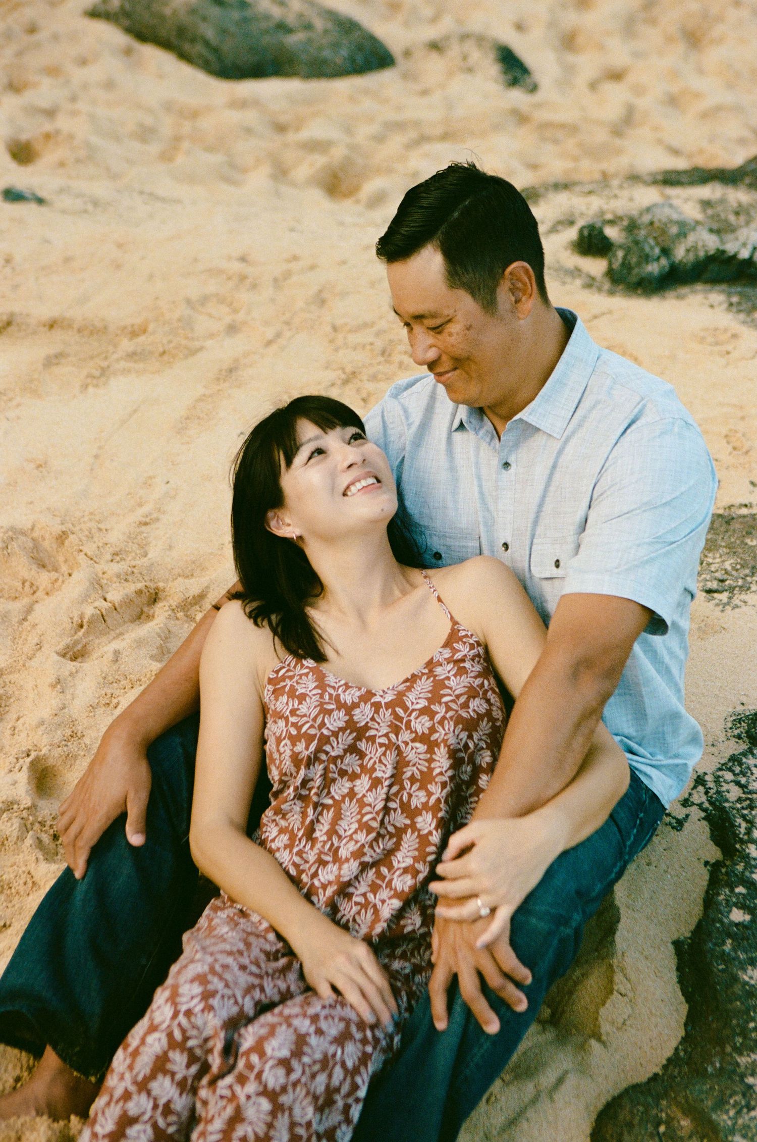 Happy Asian couple sitting together on sandy beach, woman in floral dress, man smiling warmly.