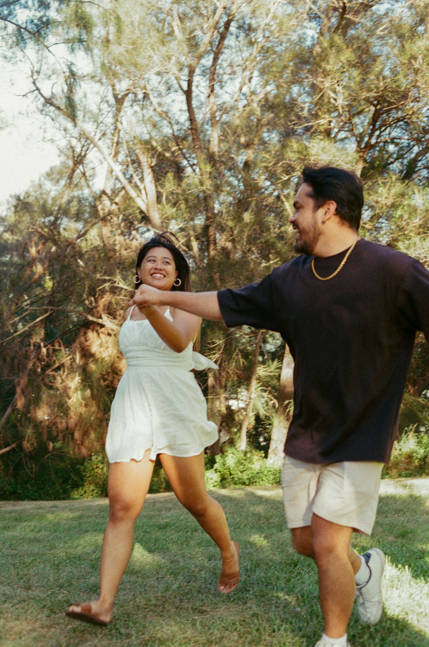 A joyful couple dances and laughs together in a sunlit park surrounded by tall trees.