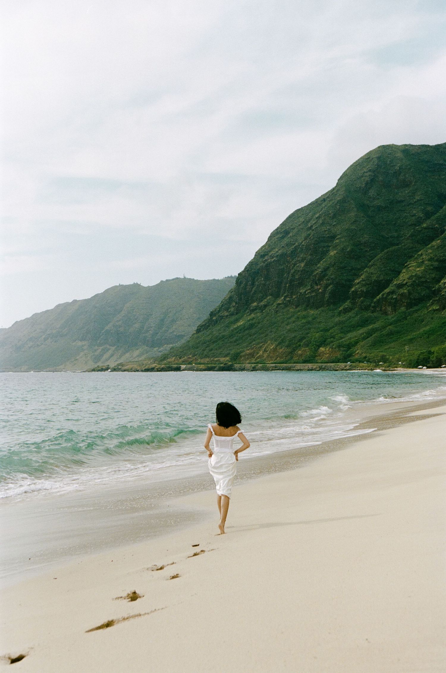 Woman in white dress walking barefoot on sandy beach leaving footprints with green mountains ahead.