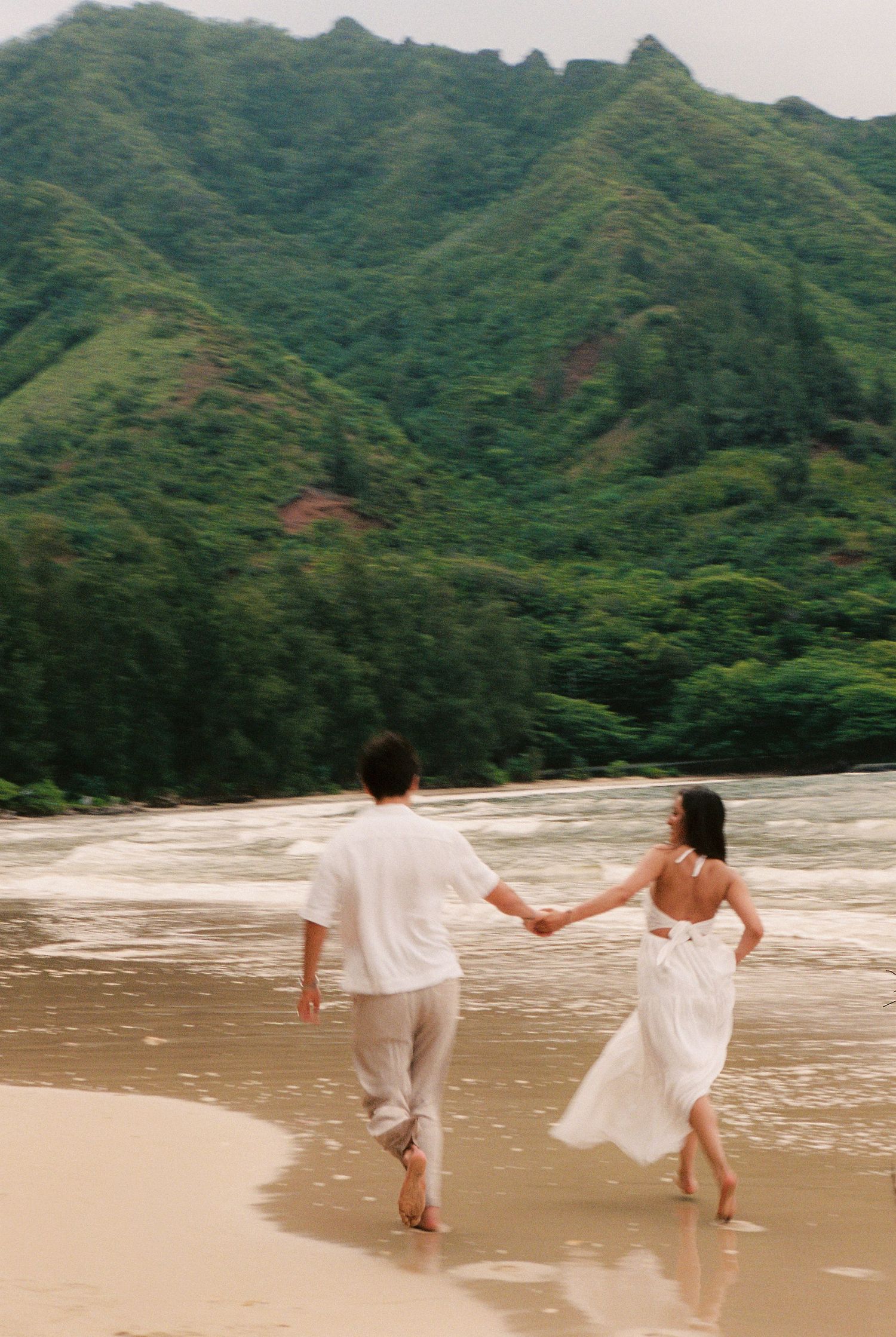 Couple in white holding hands running on a tropical beach with lush green mountains in the background.
