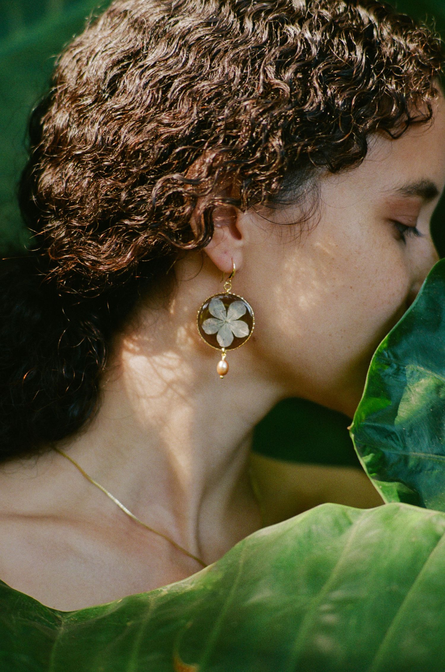 Close-up of woman with curly hair wearing gold flower and pearl drop earring near tropical leaf.