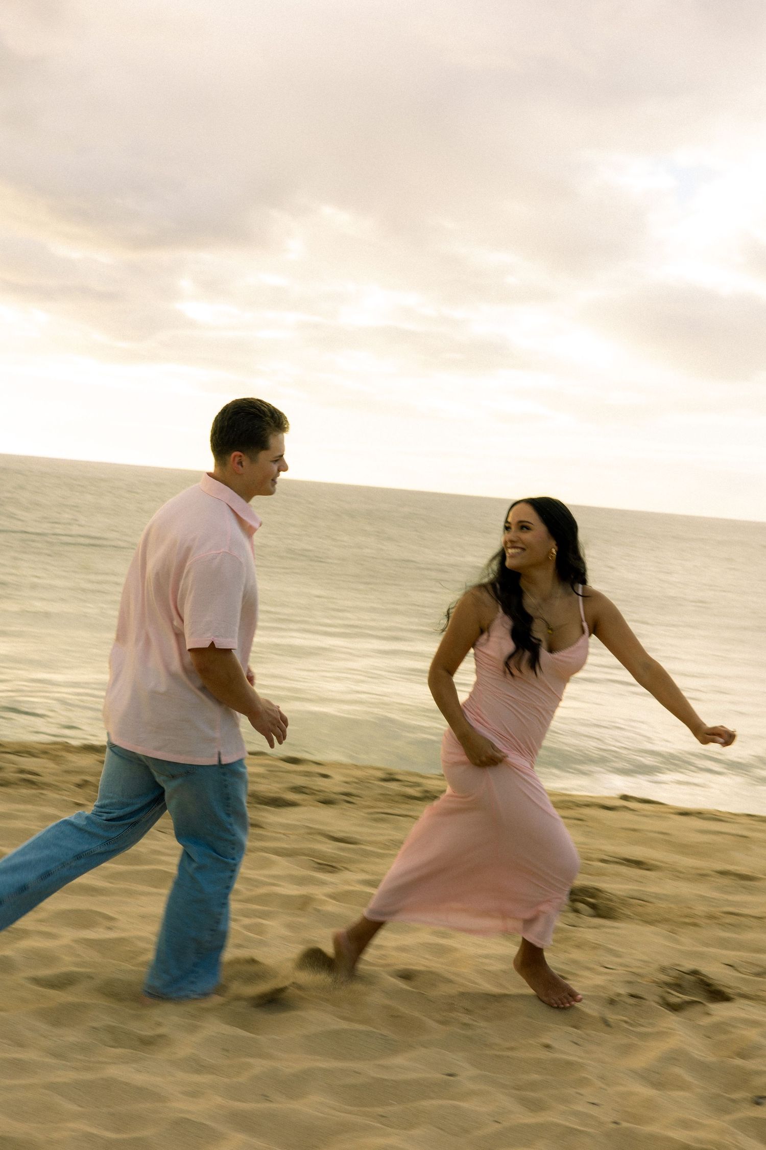 Happy couple running playfully on a sandy beach at sunset, woman wearing a flowing pink dress.