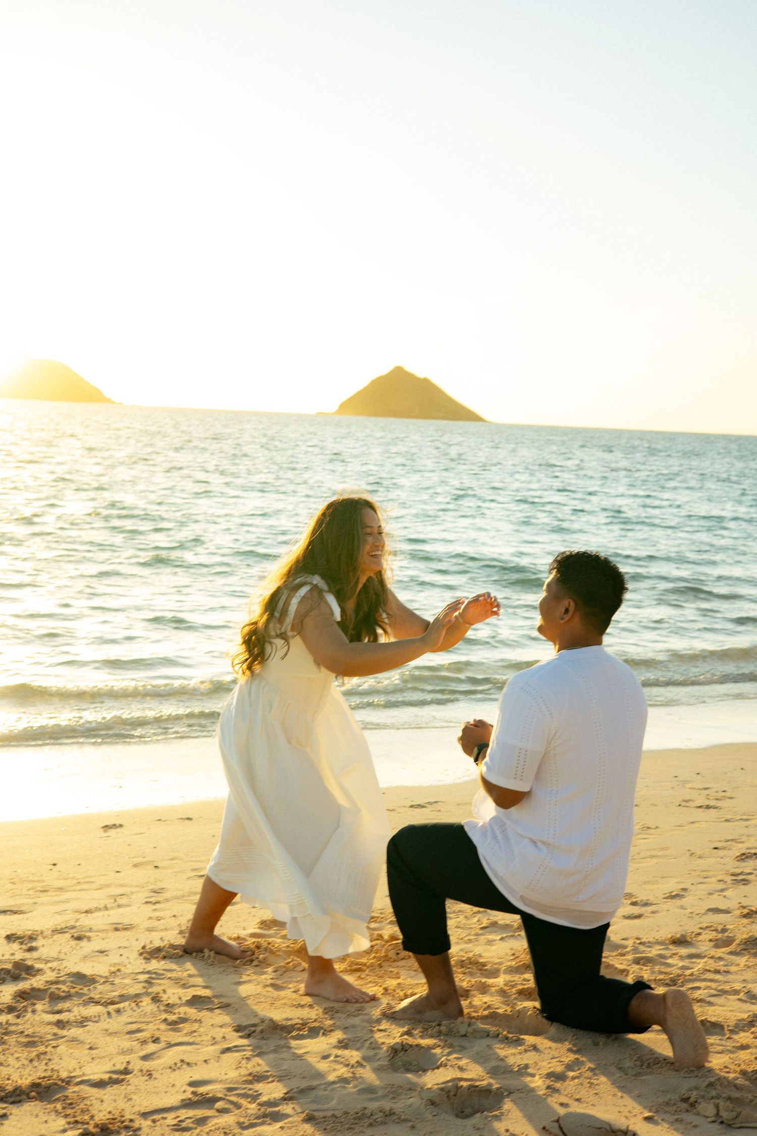 Man proposing on one knee to woman in white dress on a golden sunset beach in Hawaii.
