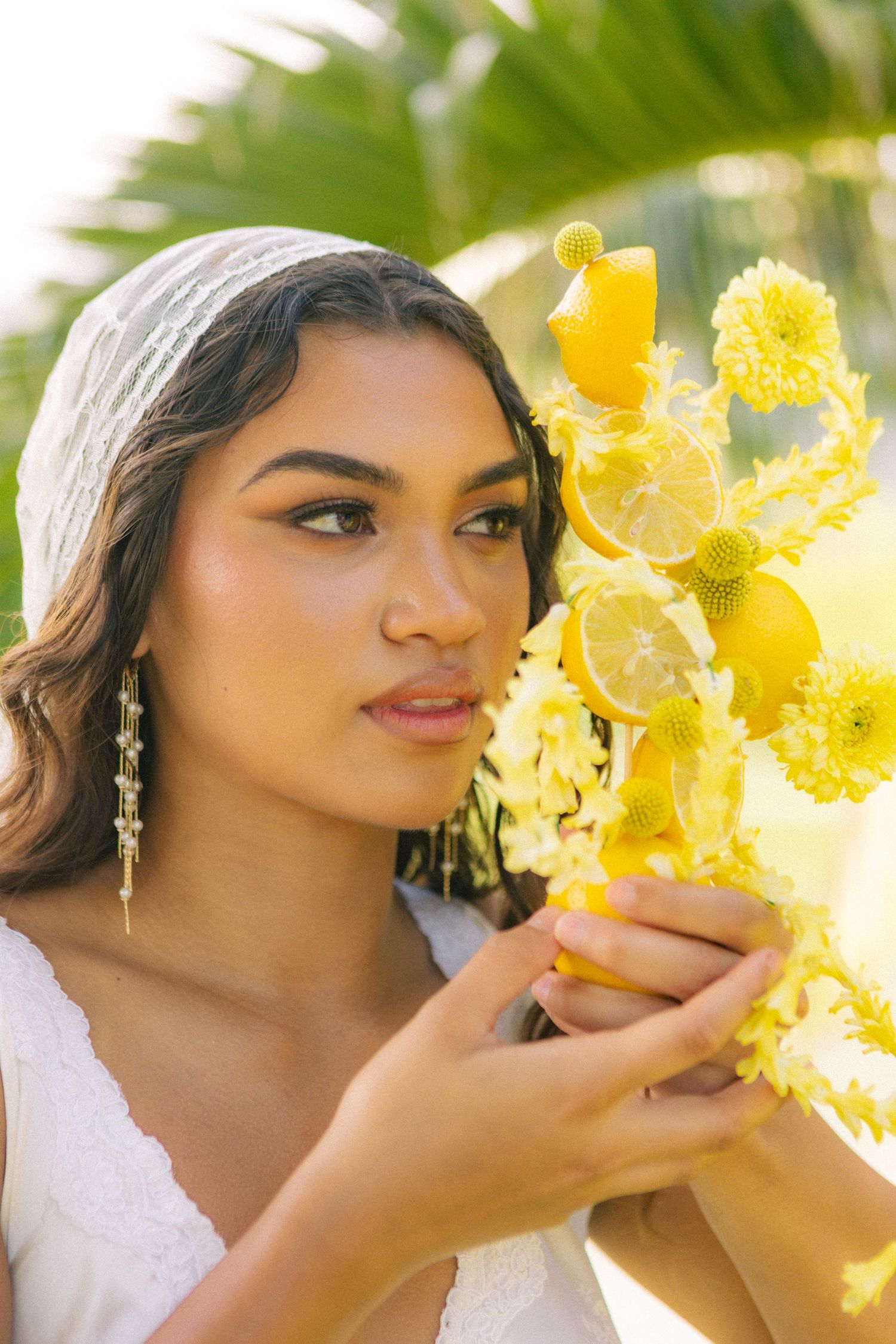 A young woman in a white dress holds a vibrant yellow bouquet of lemons and flowers, posing outdoors near palm trees.