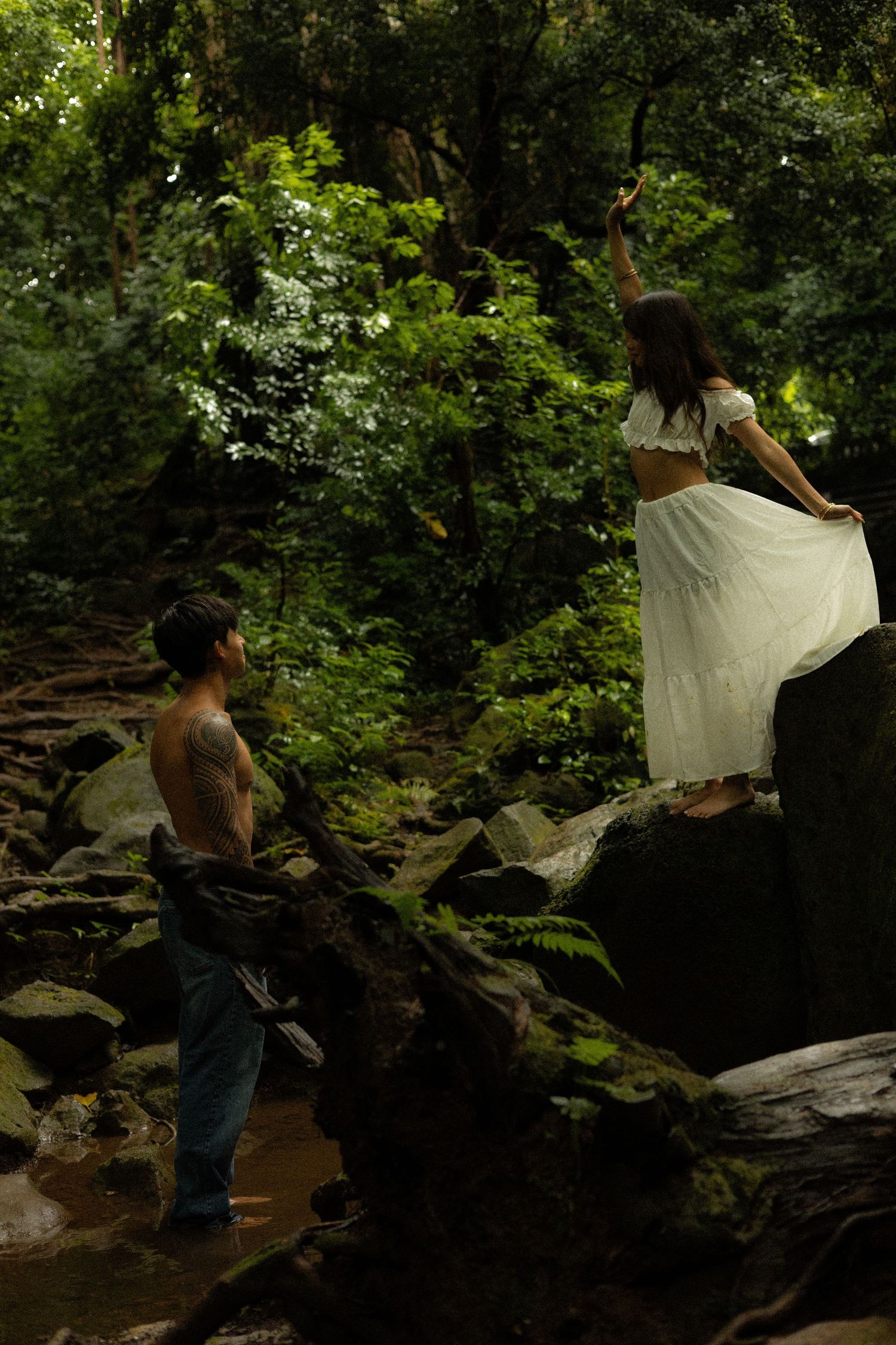 Woman in white dress standing on rocks in a lush tropical forest while man watches below.