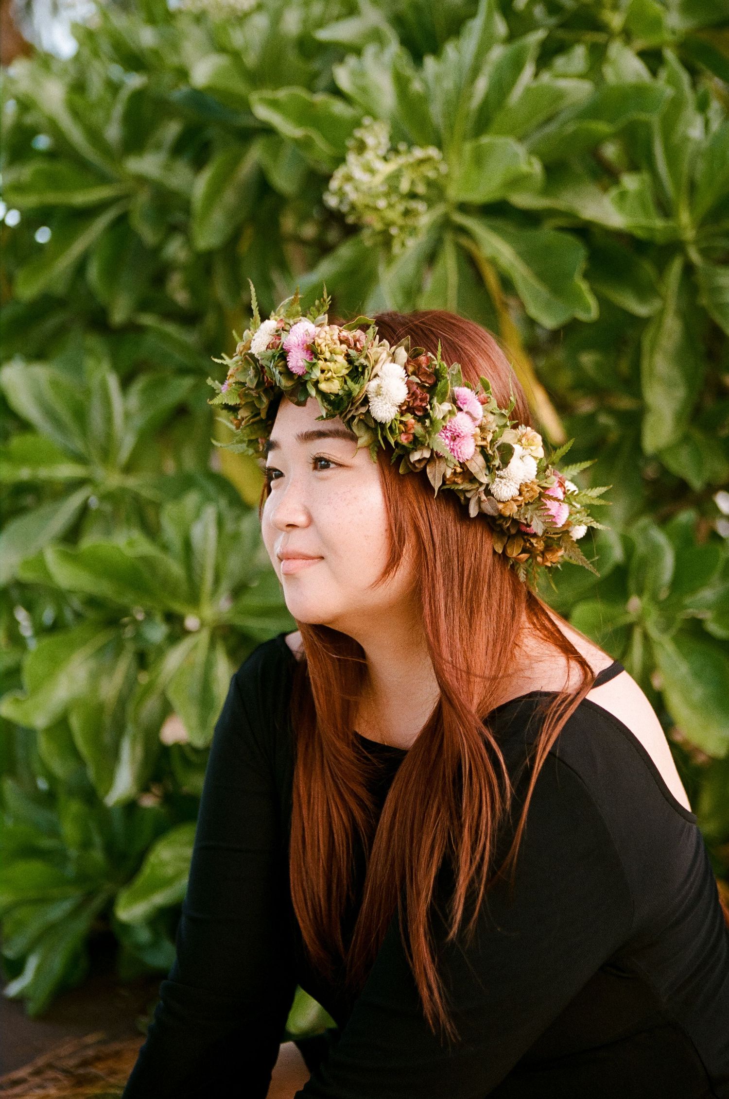 Young woman with red hair wearing floral crown sitting in front of lush green tropical foliage.
