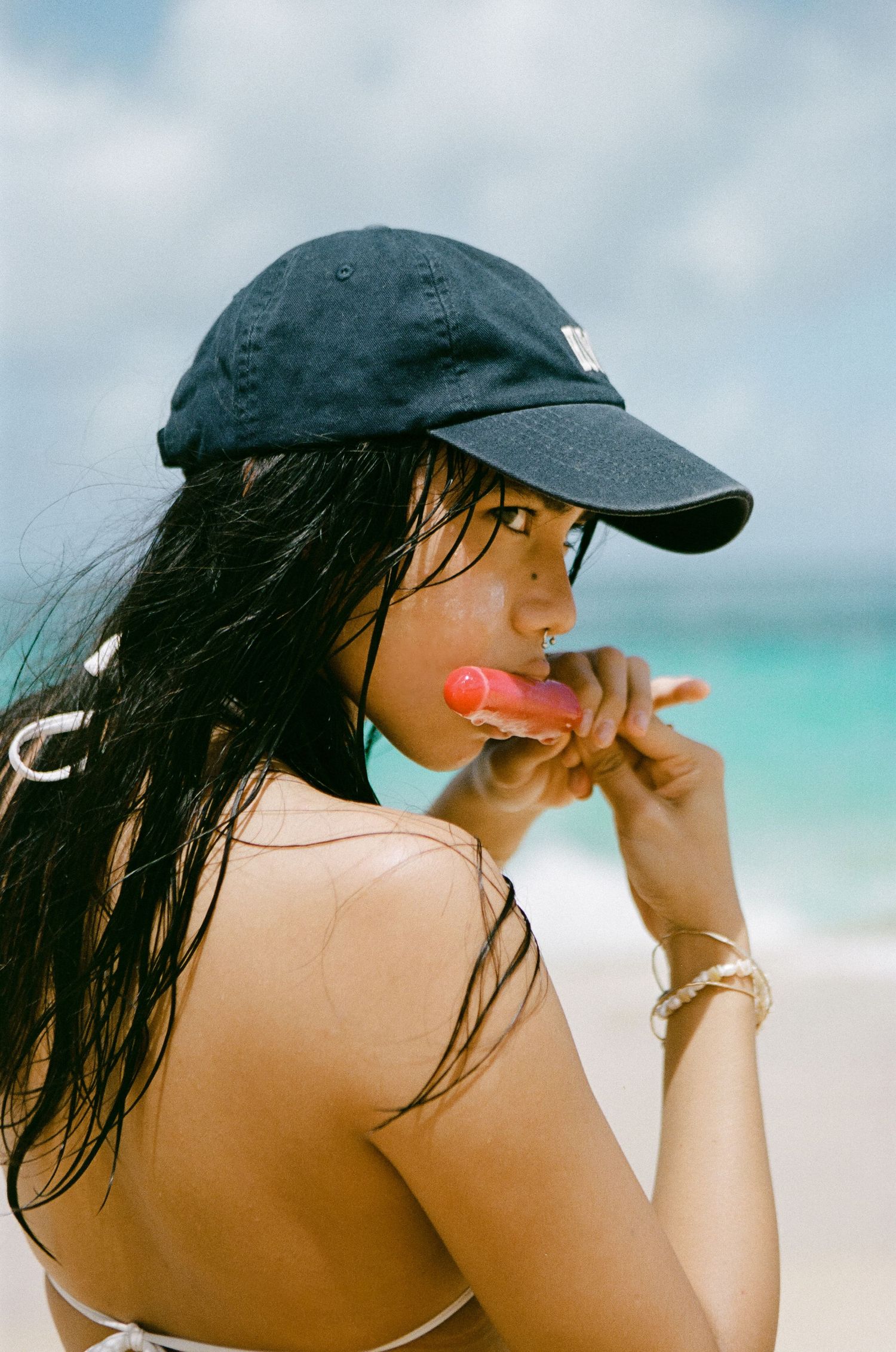 Woman in black cap eating pink popsicle on sunny beach with turquoise ocean behind her.